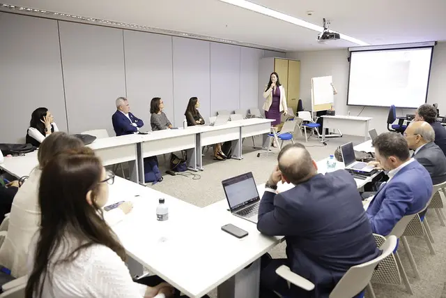 Elaine Alonso em pé, à direita, fala para um grupo de cerca de 10 pessoas sentadas em mesas brancas dispostas em formato de "U", em uma sala de aula. Ela segura um microfone e está de frente para os participantes. À direita dela, há um projetor exibindo slides e um flipchart.