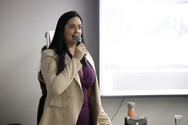 Close de Elaine Alonso, assessora do Núcleo de Sustentabilidade do TJMT, durante a oficina. Ela tem cabelos longos e pretos e usa um vestido roxo por baixo de um blazer bege aberto. Segura um microfone preto com a mão direita e está falando, olhando para a esquerda da imagem.