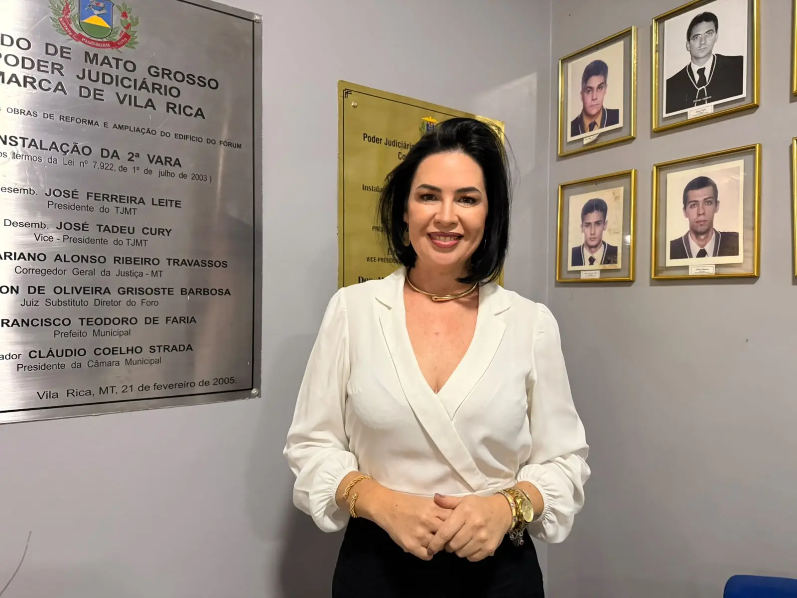 Foto horizontal que mostra a secretária de Educação de Vila Rica, Juliana Galvão, da cintura pra cima, no interior do Fórum. Ela é uma mulher branca, de cabelos pretos, lisos e com corte chanel, usa saia preta e camisa branca de manga comprida. Atrás dela, na parede, há placas de inauguração e fotos