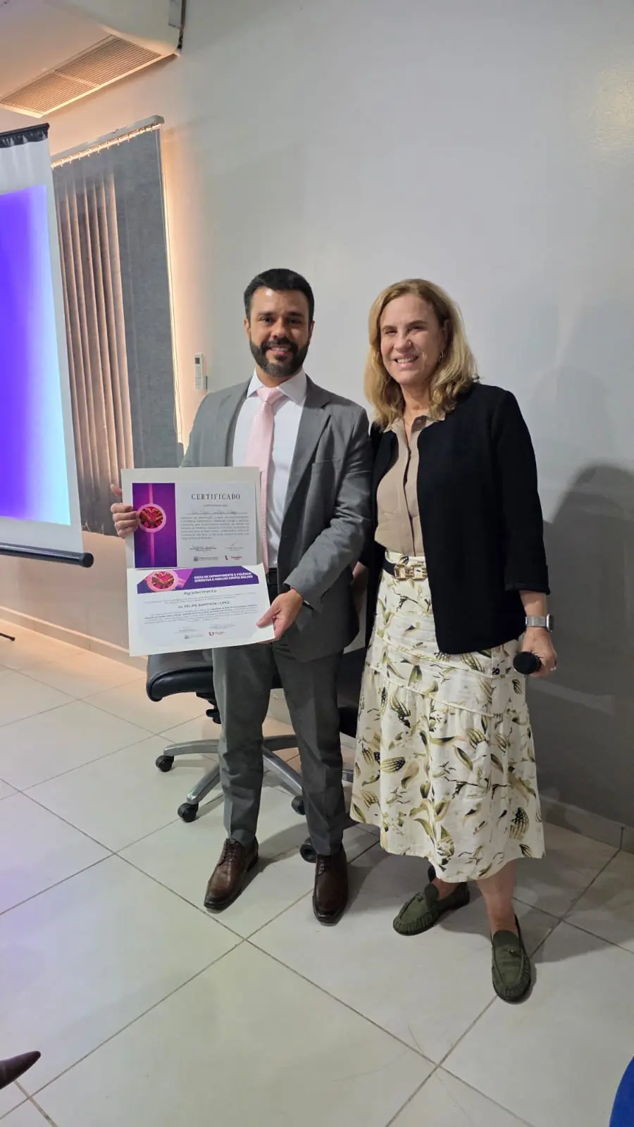 foto vertical que mostra o juiz Felipe Barthon e a desembargadora Vandymara Galvão sorrindo para a foto, em pé, no interior do fórum de Vila Rica. O juiz exibe um certificado.