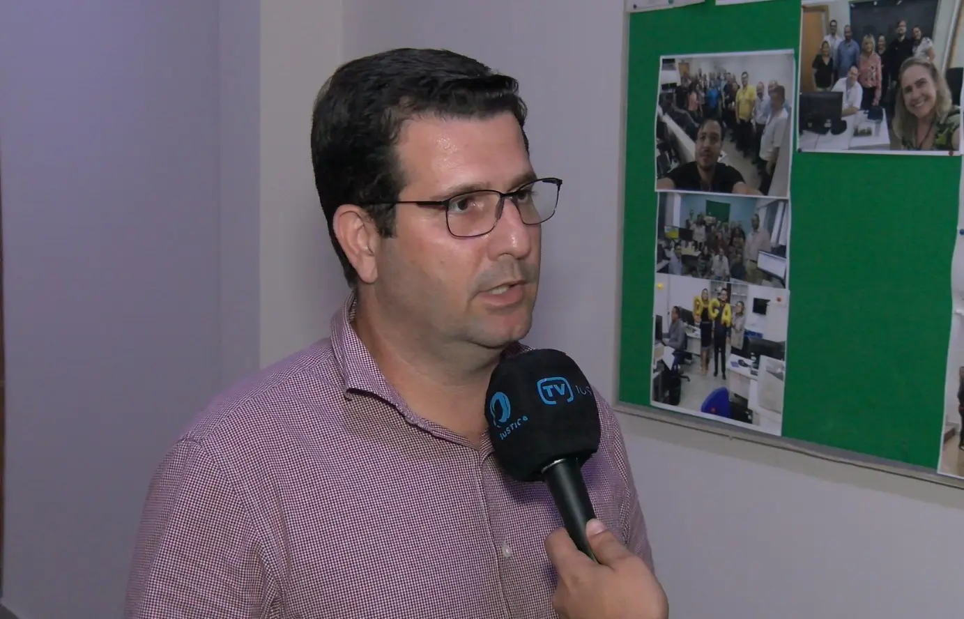 Foto horizontal em plano médio, que mostra o controlador de Arrecadação do TJMT, Daniel Vilela Balduino, durante entrevista à TV Justiça. Ele é um homem branco, de cabelos e olhos castanhos médios. Usando camisa social xadrez vermelha e branca e óculos de grau. 