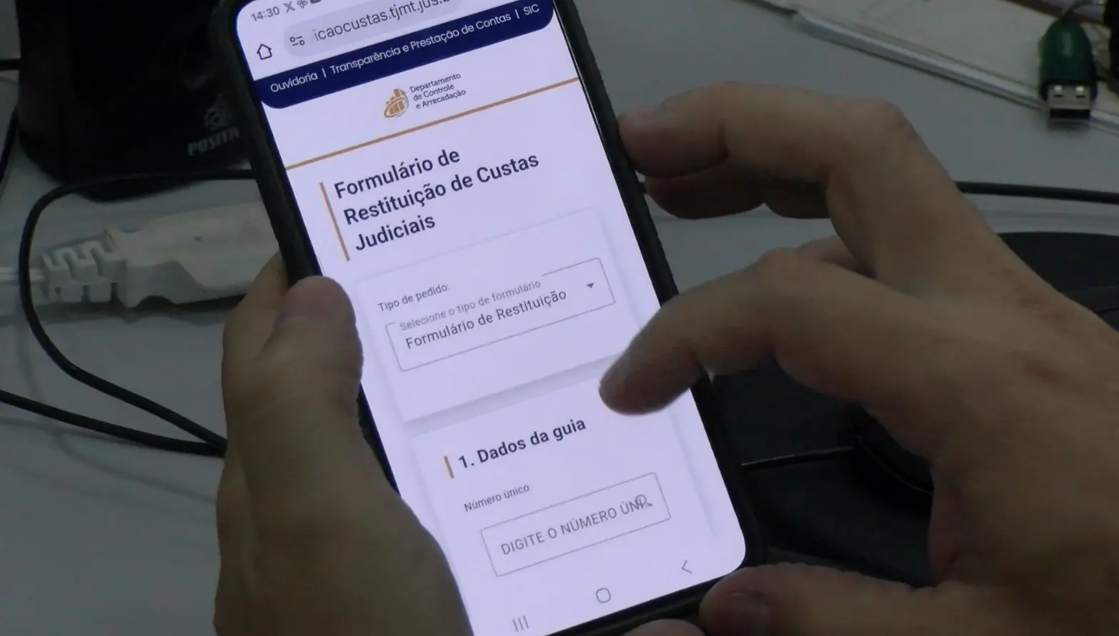 Foto horizontal em plano fechado que mostra mãos de um homem manuseando um celular, cuja tela está aberta no site do TJMT, no Formulário de Restituição de Custas Judiciais. 