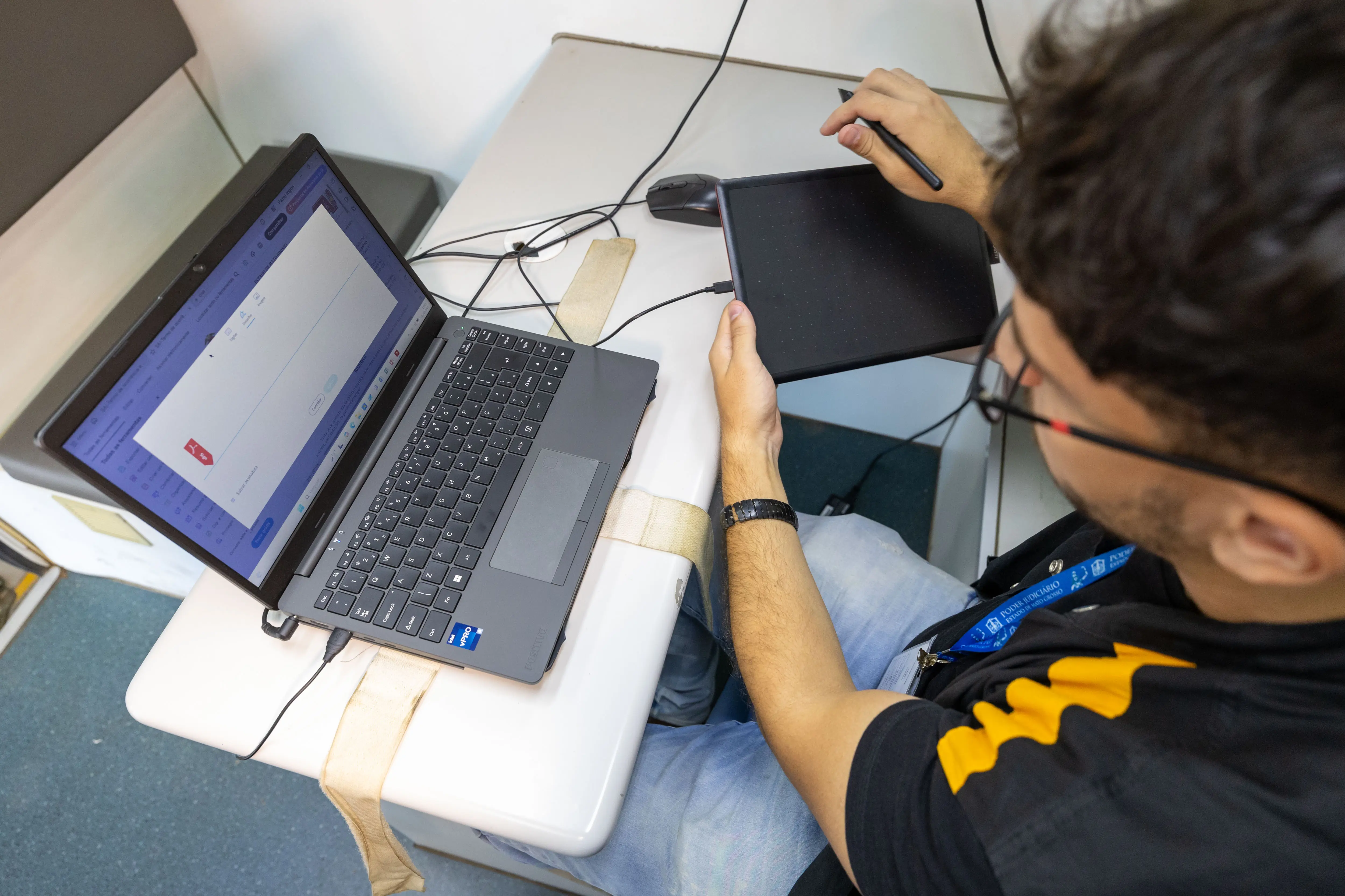 A imagem registra um homem sentado diante de um notebook, usando uma mesa digitalizadora com caneta para desenhar ou escrever. Fios conectam os equipamentos, indicando atividade tecnológica em ambiente de trabalho.