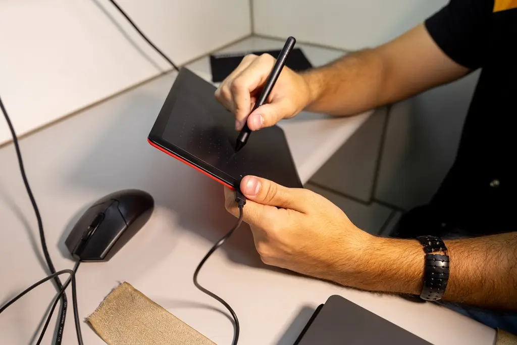 Foto horizontal em plano fechado que mostra as mãos de um homem assinando em uma mesa digitalizadora. 