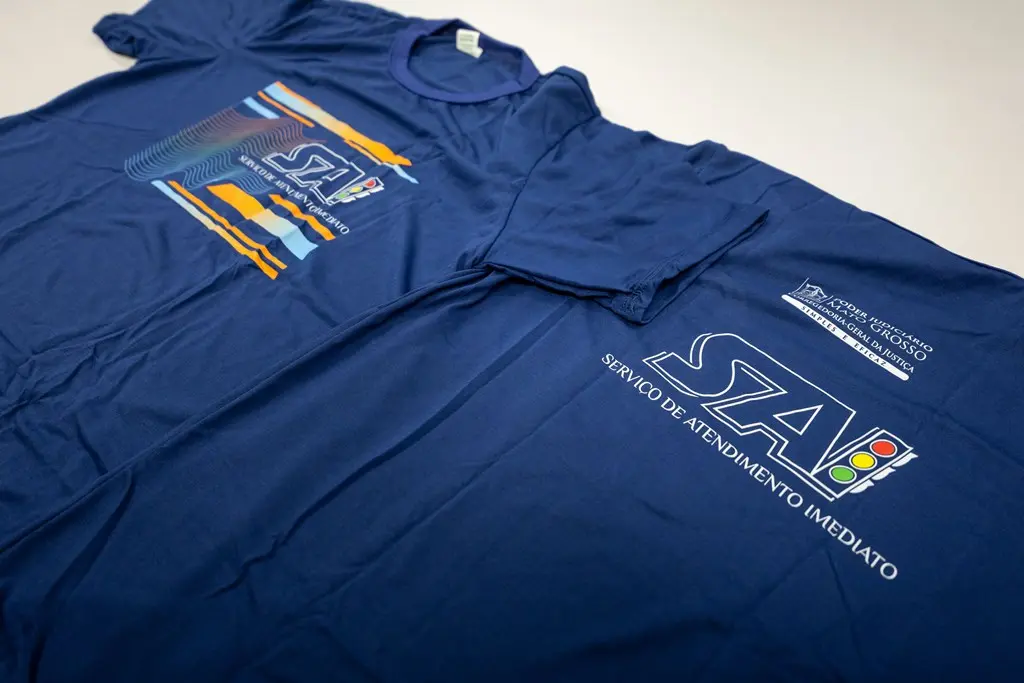 Foto horizontal em plano fechado que mostra duas camisetas do Serviço de Atendimento Imediato dispostas sobre uma mesa, uma de frente e a outra de costas, para mostrar o novo uniforme do SAI. A camiseta é azul marinho e tem a logomarca do SAI de forma estilizada na frente e tradicional nas costas. 