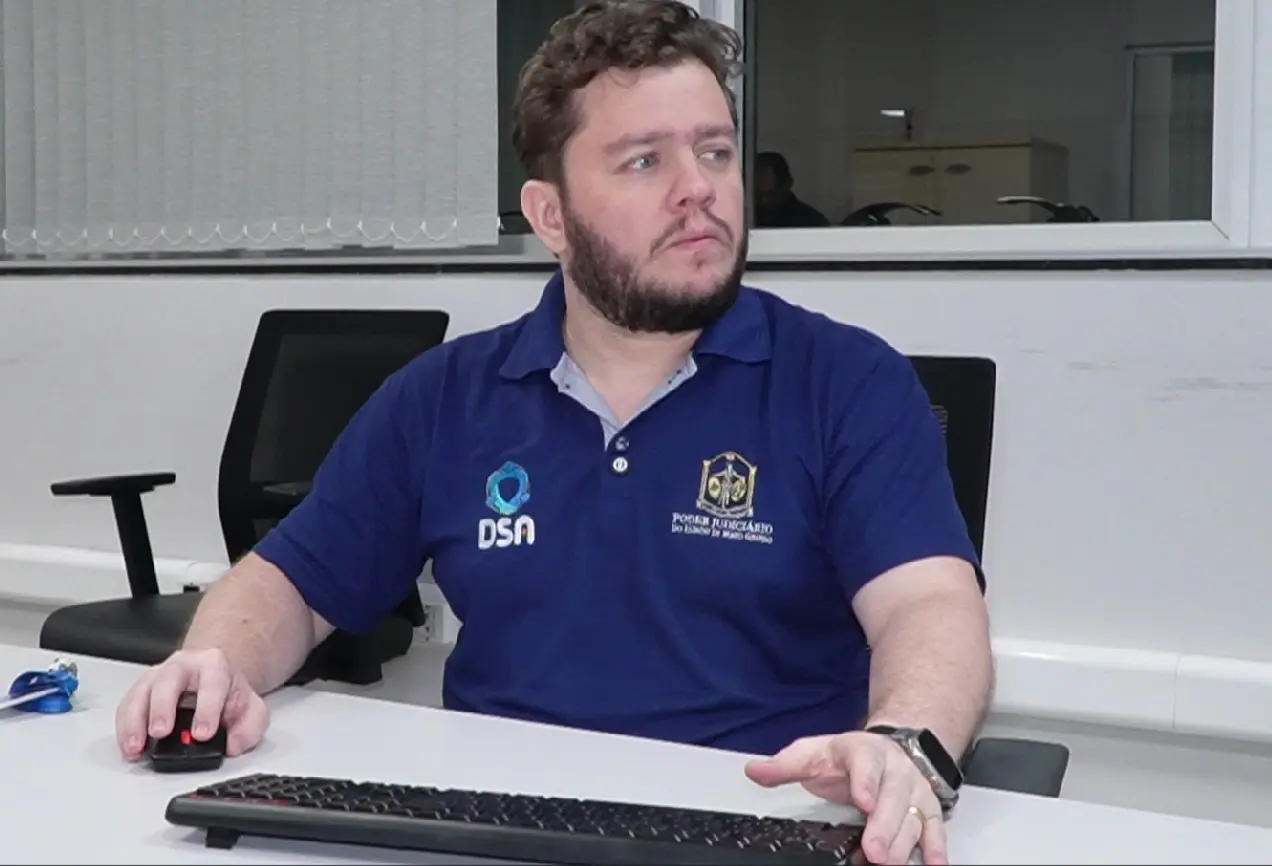 Um homem de cabelos castanhos e barba curta veste polo azul com emblemas do Poder Judiciário de Mato Grosso e da DSA. Está sentado à mesa com teclado e mouse.
