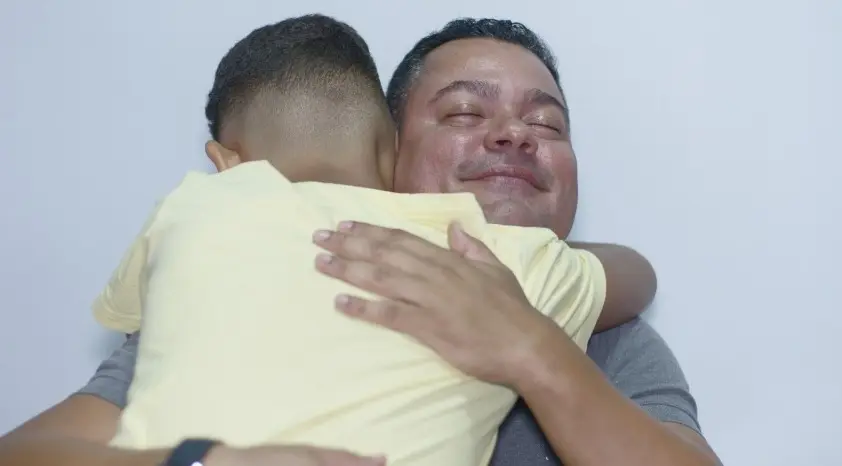Imagem mostra padrinho e afilhado em um abraço. O Padrinho está em destaque. Ele é um homem pardo e está sorrindo. A criança está de costas e veste uma camisa amarela.