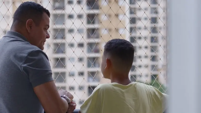 Imagem mostra um homem e uma criança conversando em uma sacada de prédio. O homem veste uma camiseta cinza e o garoto uma blusa amarela.