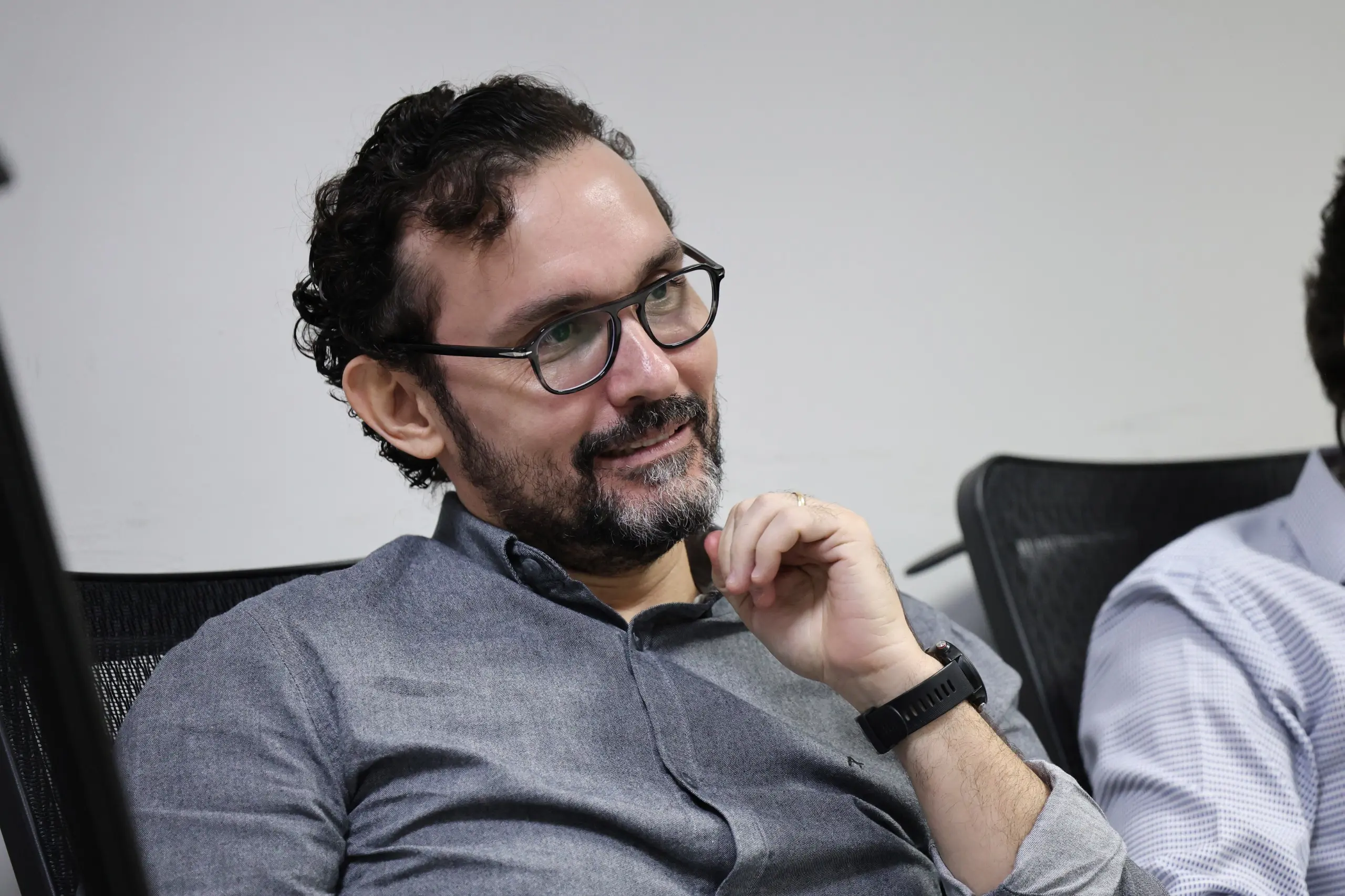 Homem sentado em cadeira de sala de aula ou auditório, com postura atenta e levemente inclinada para trás. Usa camisa de manga longa em tom escuro e relógio no pulso. Uma das mãos está próxima ao queixo, em gesto de reflexão, enquanto ele acompanha a atividade. 