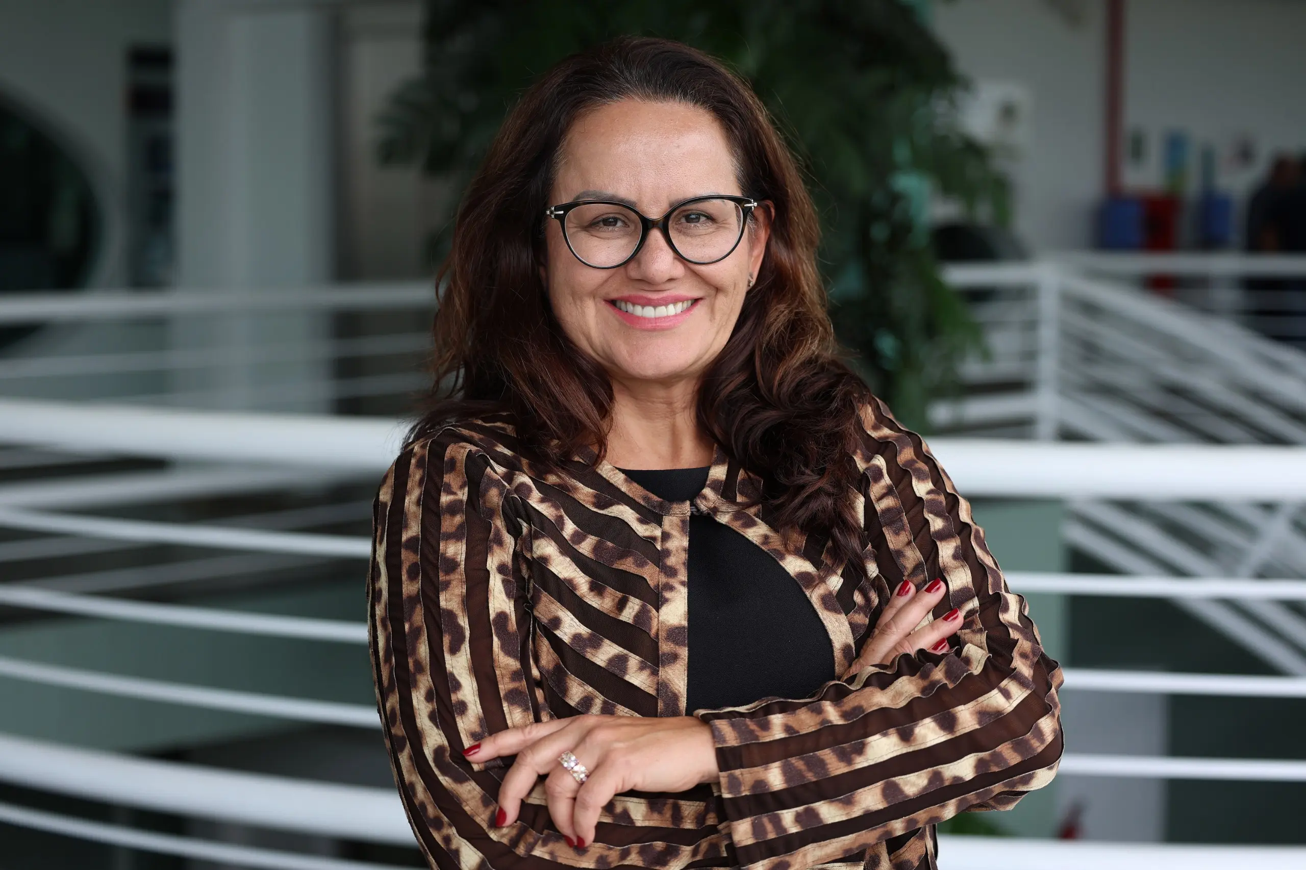 Mulher em primeiro plano, em ambiente de sala de aula, usando blusa com estampa em tons terrosos e cabelos soltos na altura dos ombros. Ao fundo, aparecem outros participantes sentados e um quadro branco, indicando um contexto acadêmico ou de formação