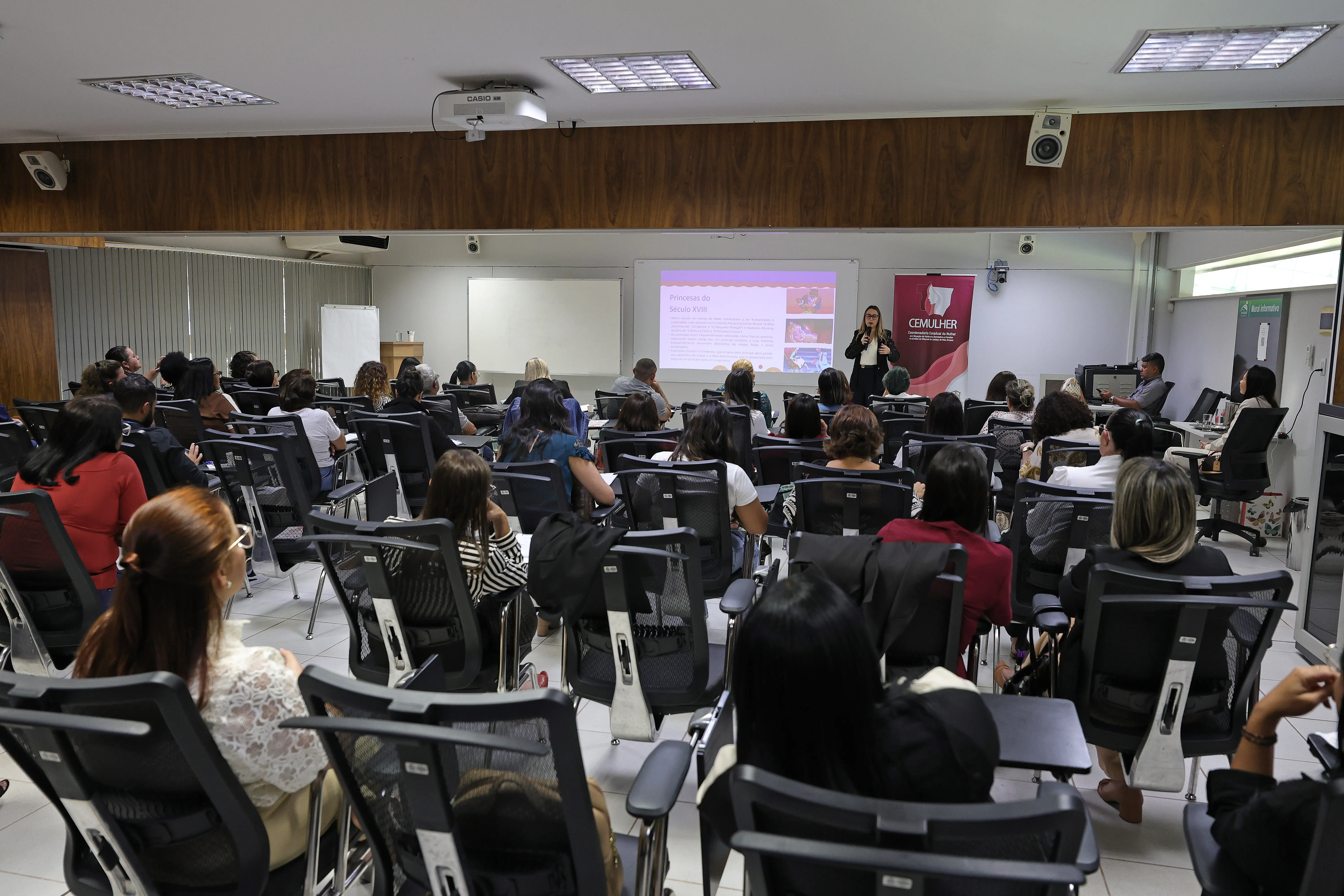 Plateia sentada assiste palestra em auditório. Ao fundo, palestrante de preto fala ao microfone diante de telão com slide e banner do CEMULHER.