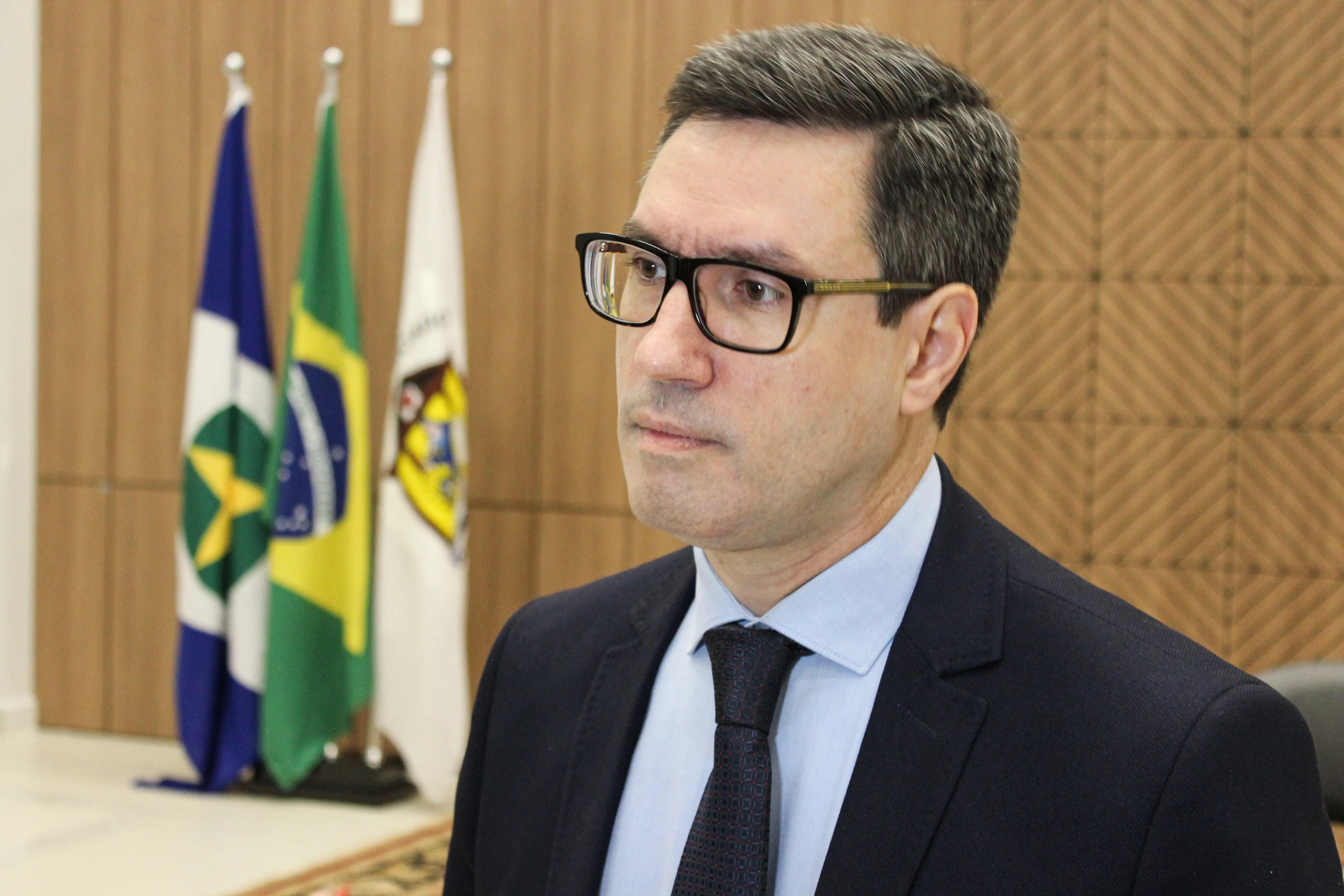 Homem de cabelos curtos escuros e óculos de armação preta enquadrado em plano médio curto. Ele veste paletó escuro, camisa azul-claro e gravata escura. Ao fundo, veem-se três bandeiras hasteadas e painel de madeira.