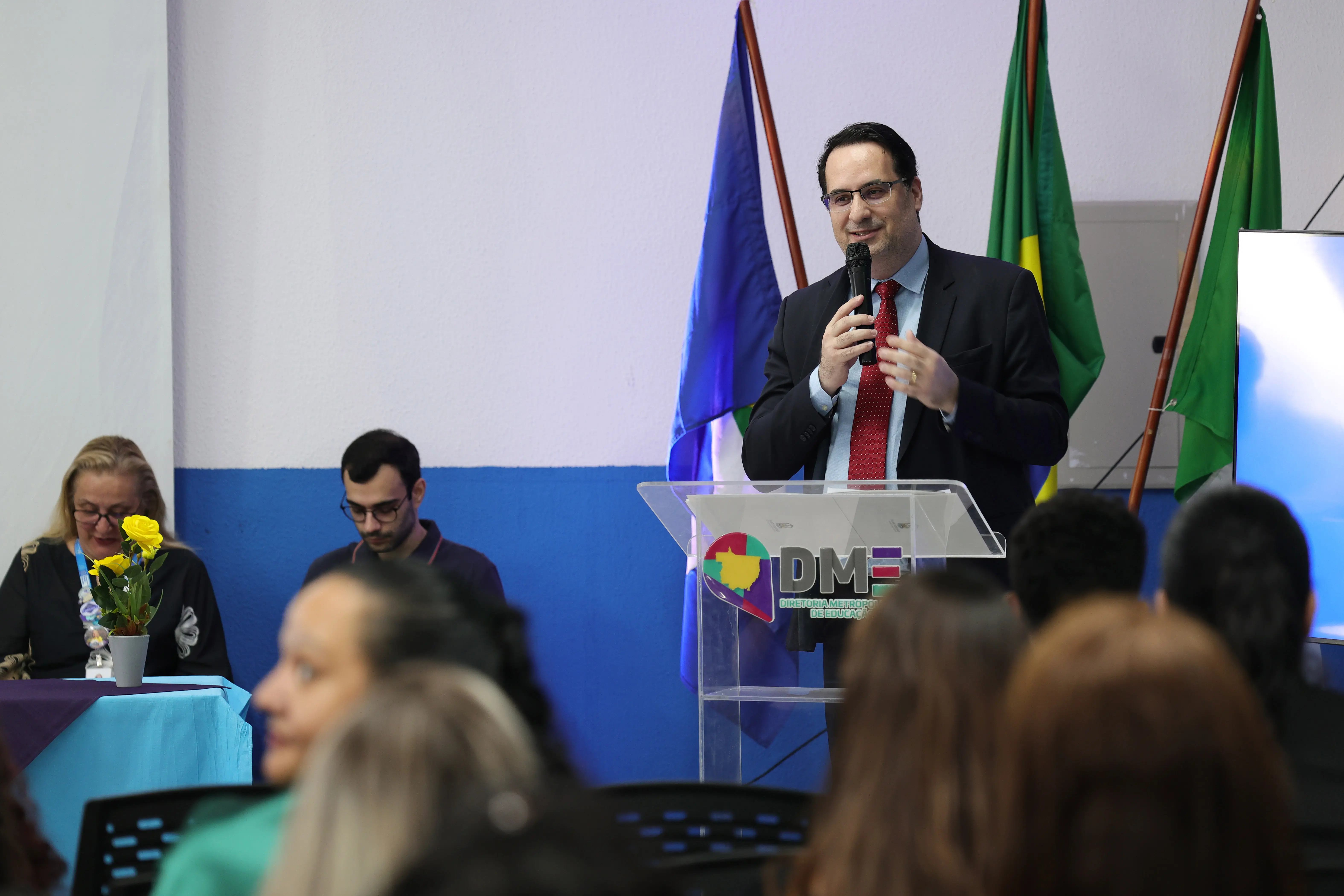 Homem de terno escuro, camisa azul-clara e gravata vermelha, óculos, fala ao microfone em púlpito com o logotipo da Diretoria Metropolitana de Educação. Ao fundo, bandeiras estaduais e do Brasil.