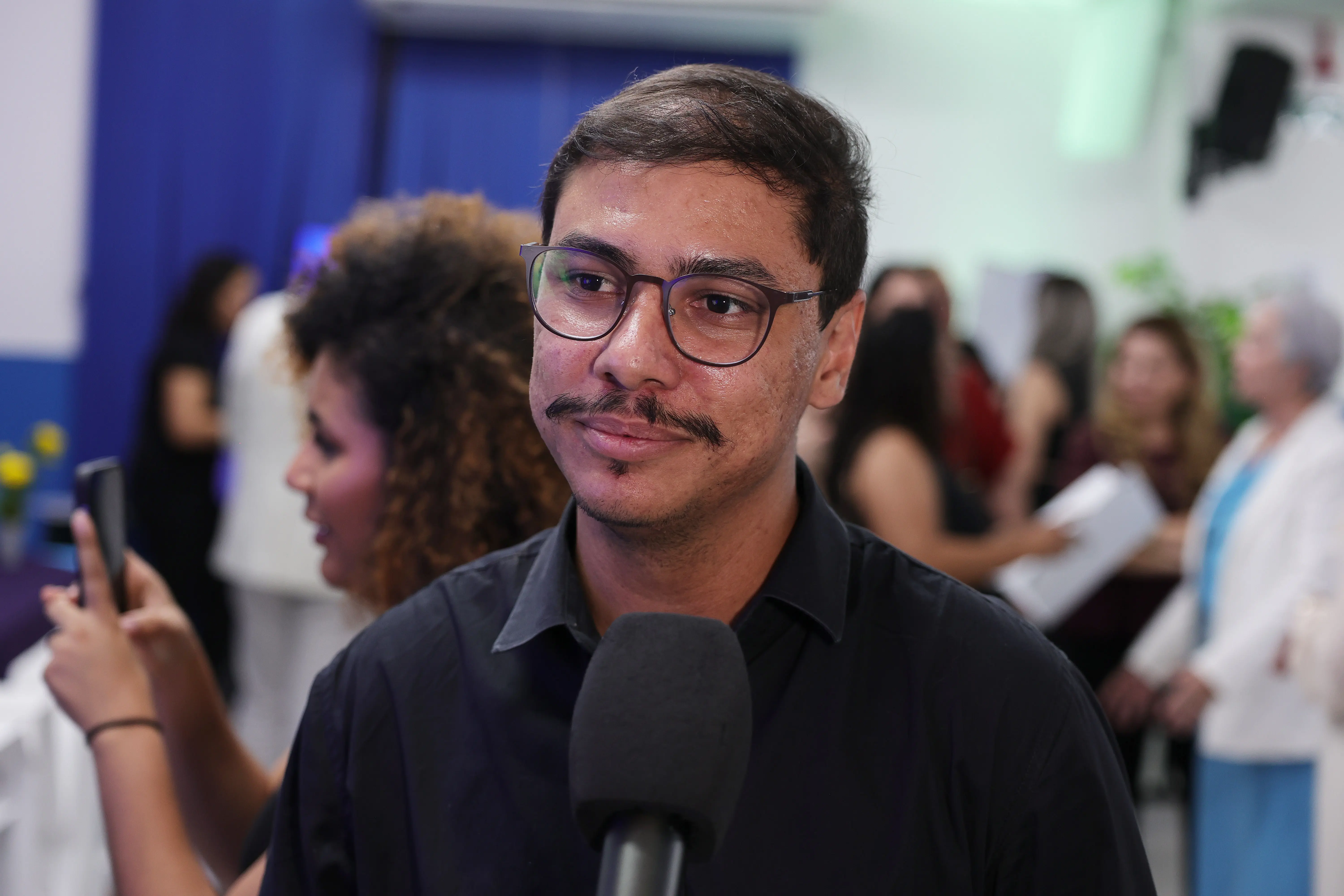 Homem jovem, óculos de grau e bigode, veste camisa preta, aparece próximo a um microfone durante evento. Ao fundo, participantes registram o momento com celular.
