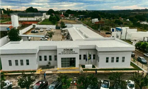 Vista aérea da fachada do Fórum de Alto Araguaia, um prédio térreo branco com três bandeiras na entrada. Há carros estacionados ao redor e uma área residencial e arborizada ao fundo.