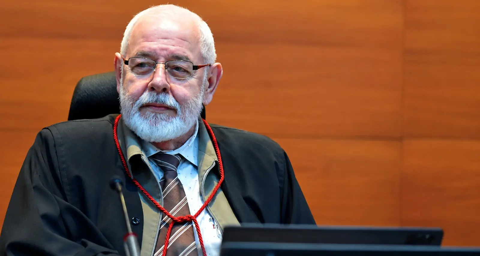 Foto horizontal que mostra o presidente do TJMT, desembargador José Zuquim Nogueira, sentado em seu posto no plenário do tribunal, olhando para o lado. Ele é um senhor branco, de cabelo e barba brancos, usando toga e óculos de grau.