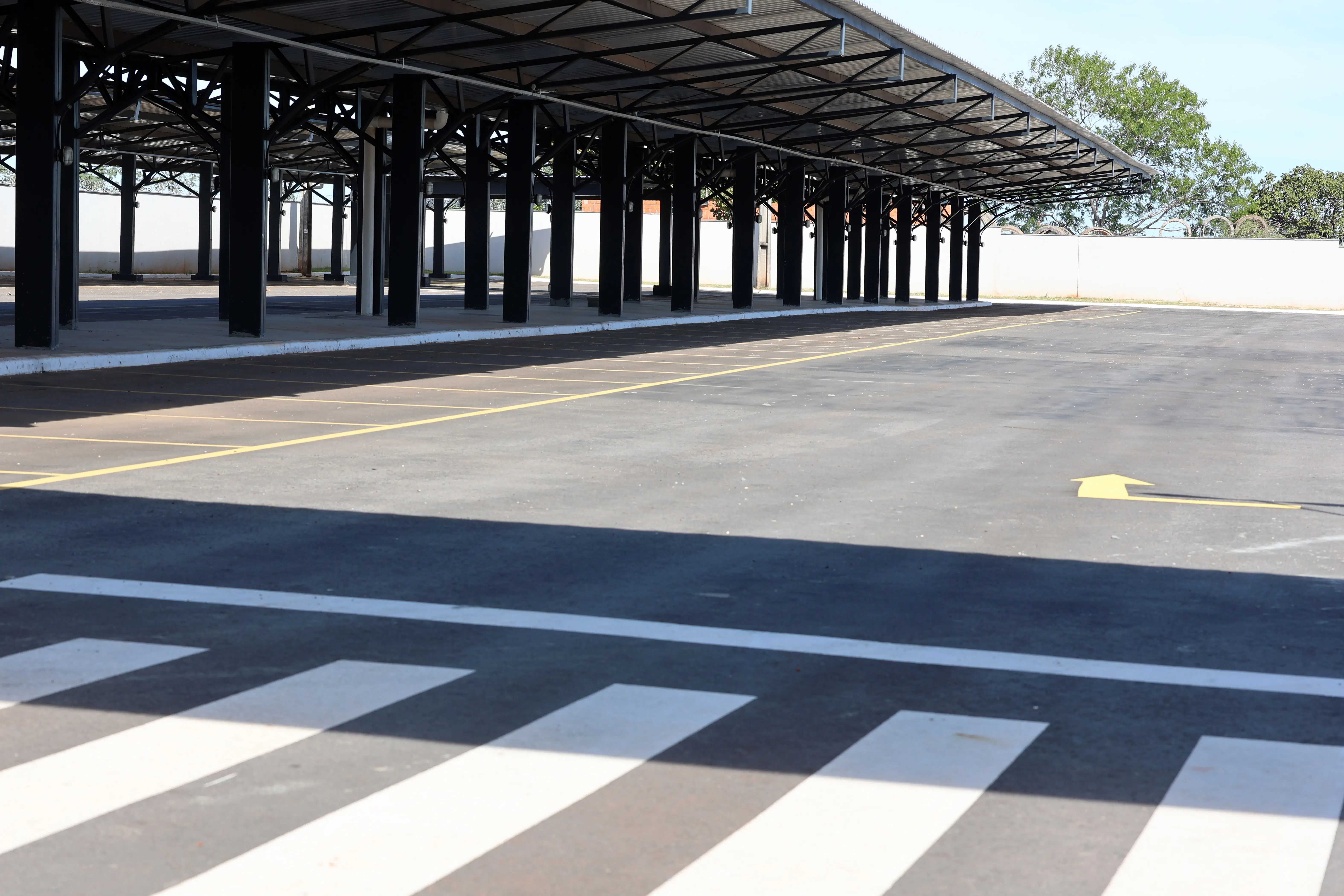Estacionamento amplo e vazio, com asfalto novo e vagas demarcadas por linhas amarelas. À esquerda, uma grande estrutura de cobertura metálica com pilares pretos. Em primeiro plano, uma faixa de pedestres.