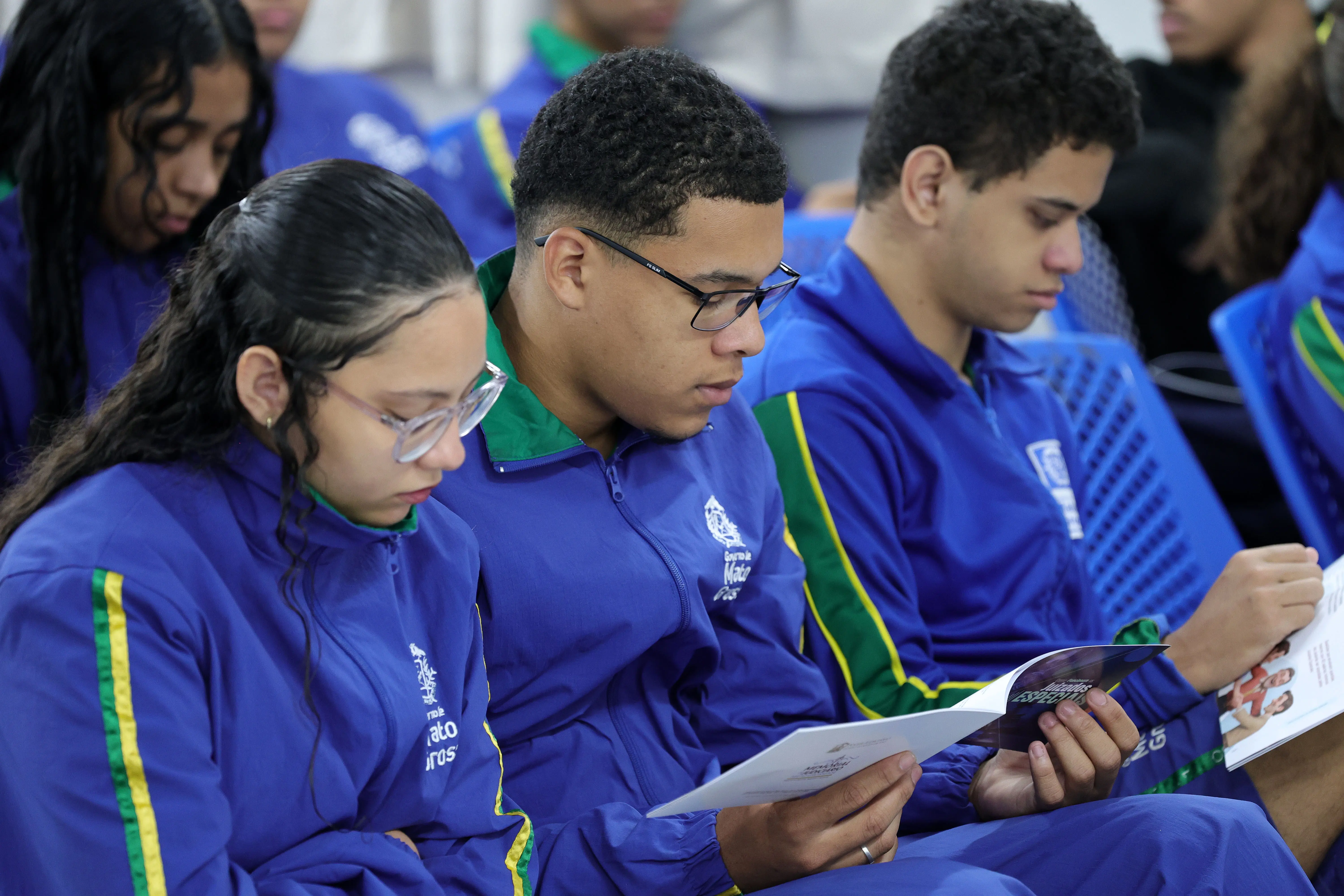 Estudantes sentados, usando uniformes azuis com faixas verdes e amarelas, leem folhetos intitulados 