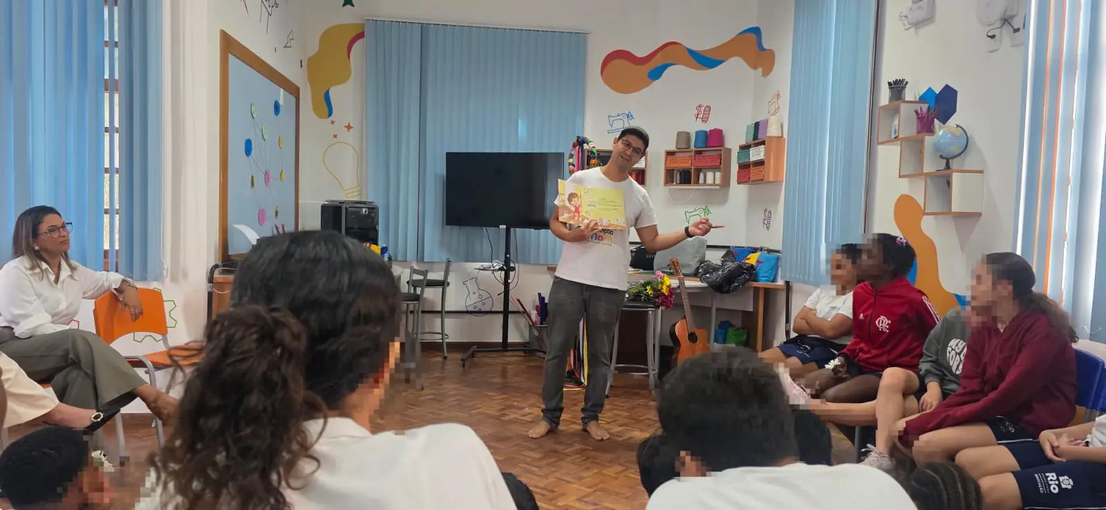 Em uma sala de aula decorada com desenhos coloridos, um homem de camiseta branca segura um livro aberto e aponta para o lado enquanto fala. Várias pessoas e jovens estão sentados ao redor ouvindo a apresentação.