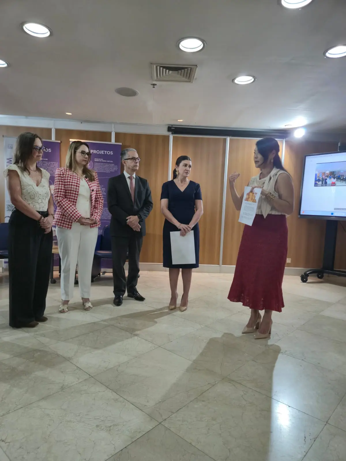 Uma mulher de saia vermelha e blusa clara segura uma folha e fala de pé para um grupo de quatro pessoas alinhadas ao seu lado — três mulheres e um homem de terno. Ao fundo, há um painel roxo com texto e uma tela de televisão.
