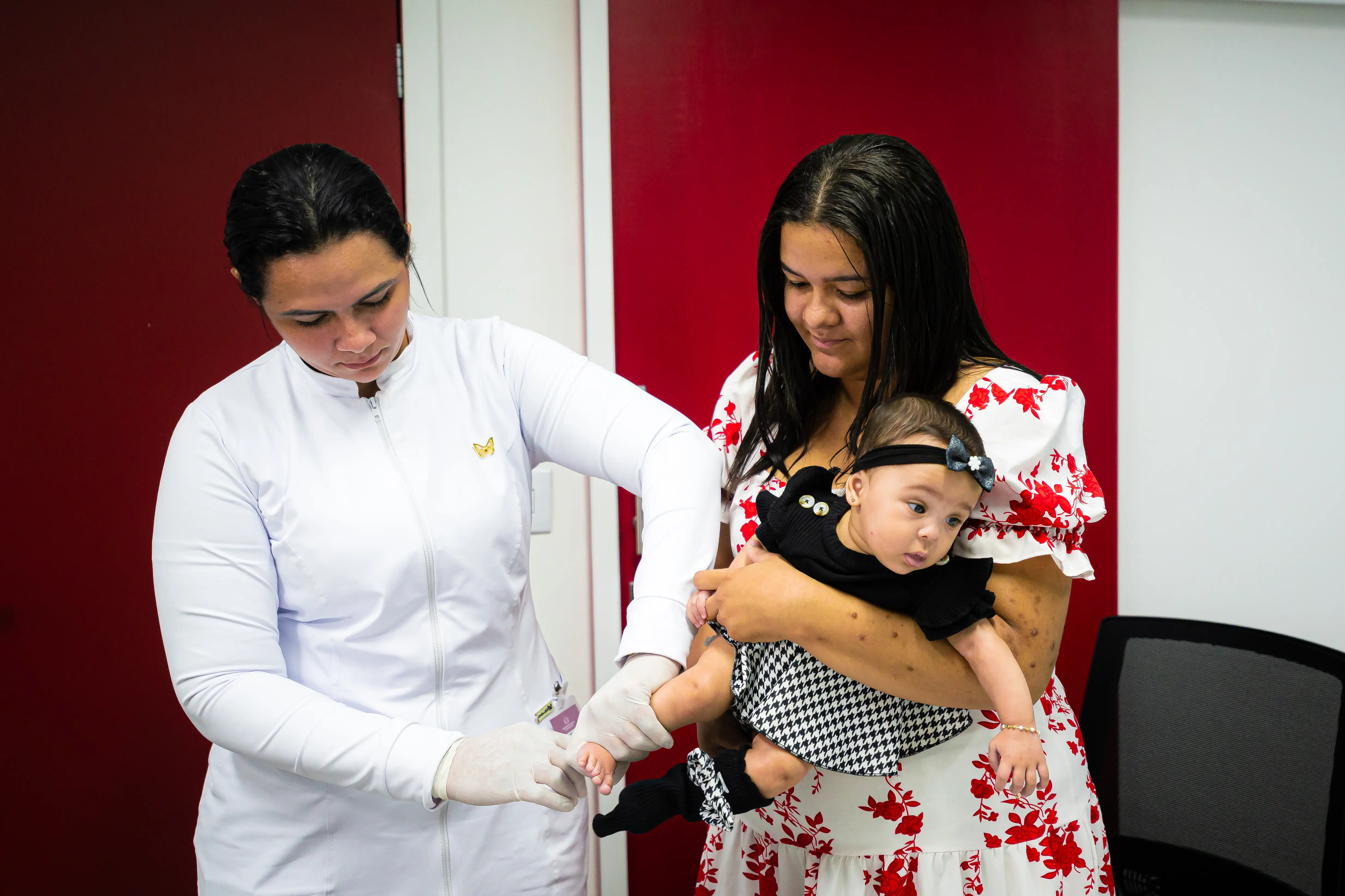 Uma profissional de branco, com luvas, examina o pezinho de um bebê. Ao lado, a mãe, de vestido floral, segura a criança no colo. Parede vermelha ao fundo.
