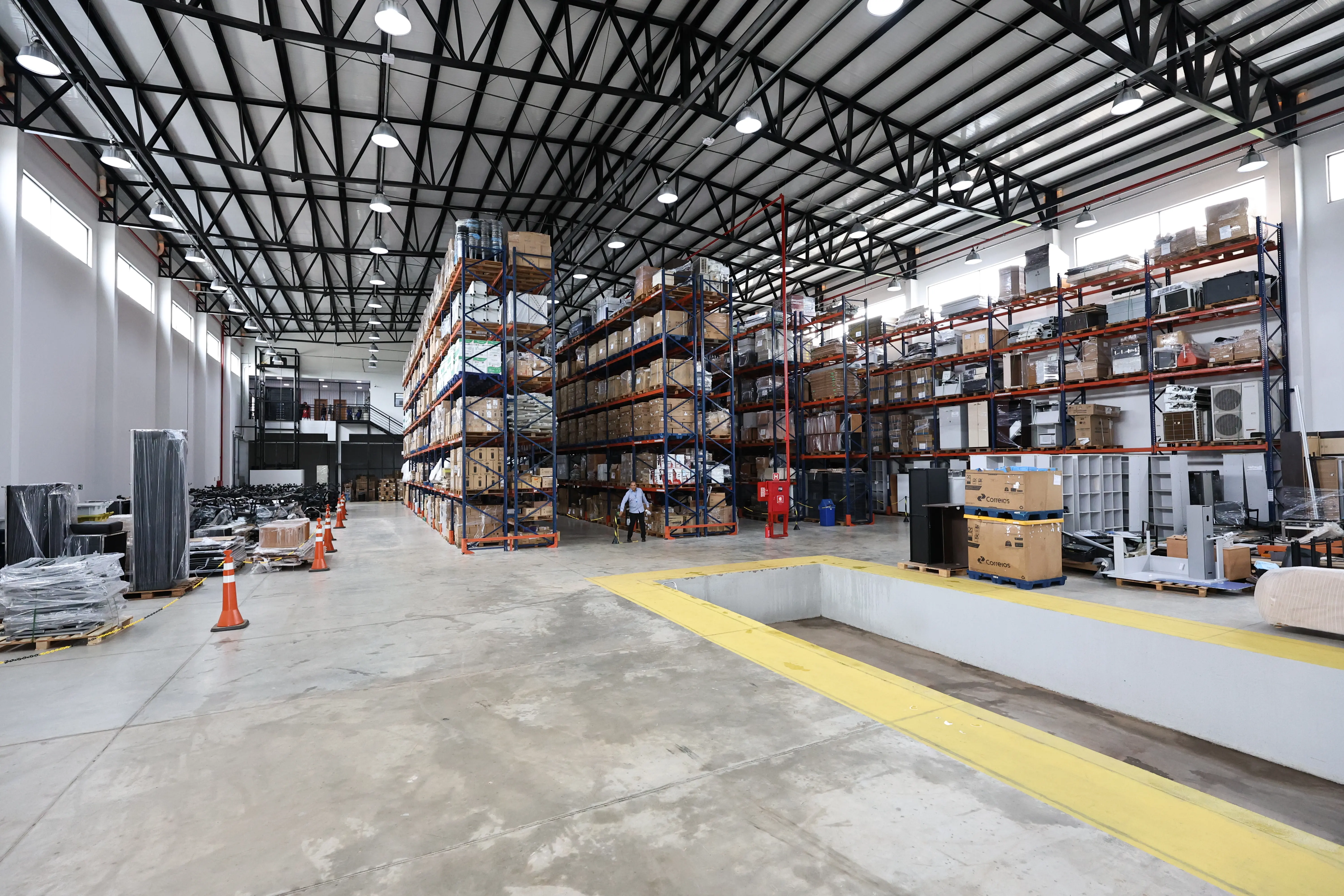 Interior de um galpão industrial ou depósito amplo com teto alto e estrutura metálica. Grandes prateleiras verticais de aço organizam caixas e caixotes de armazenamento. Chão de cimento cinza.