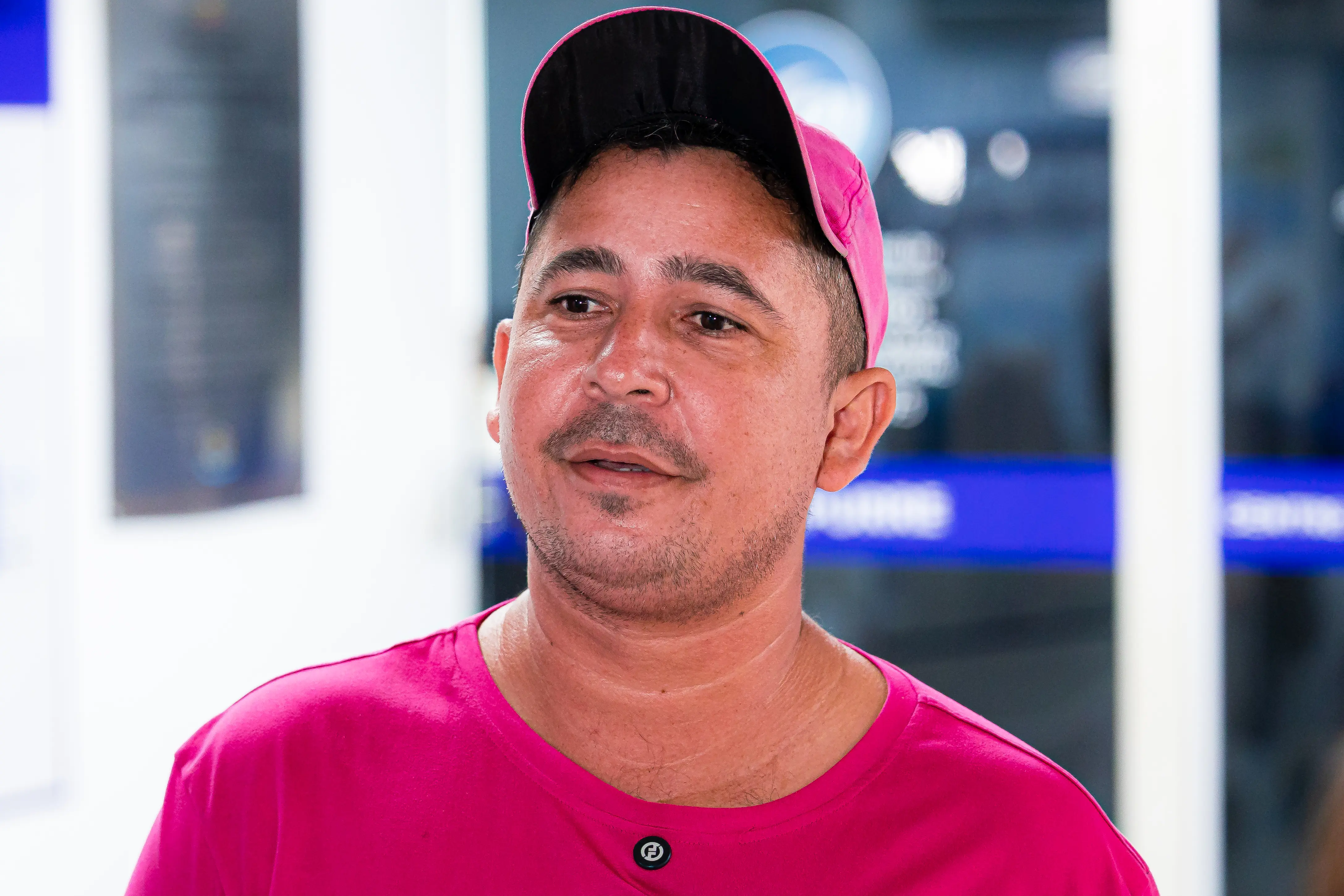 Um homem usa boné rosa e camiseta rosa, tem bigode e barba por fazer, olha para o lado e sorri levemente. Ao fundo, ambiente interno com painéis e tela azul desfocados.