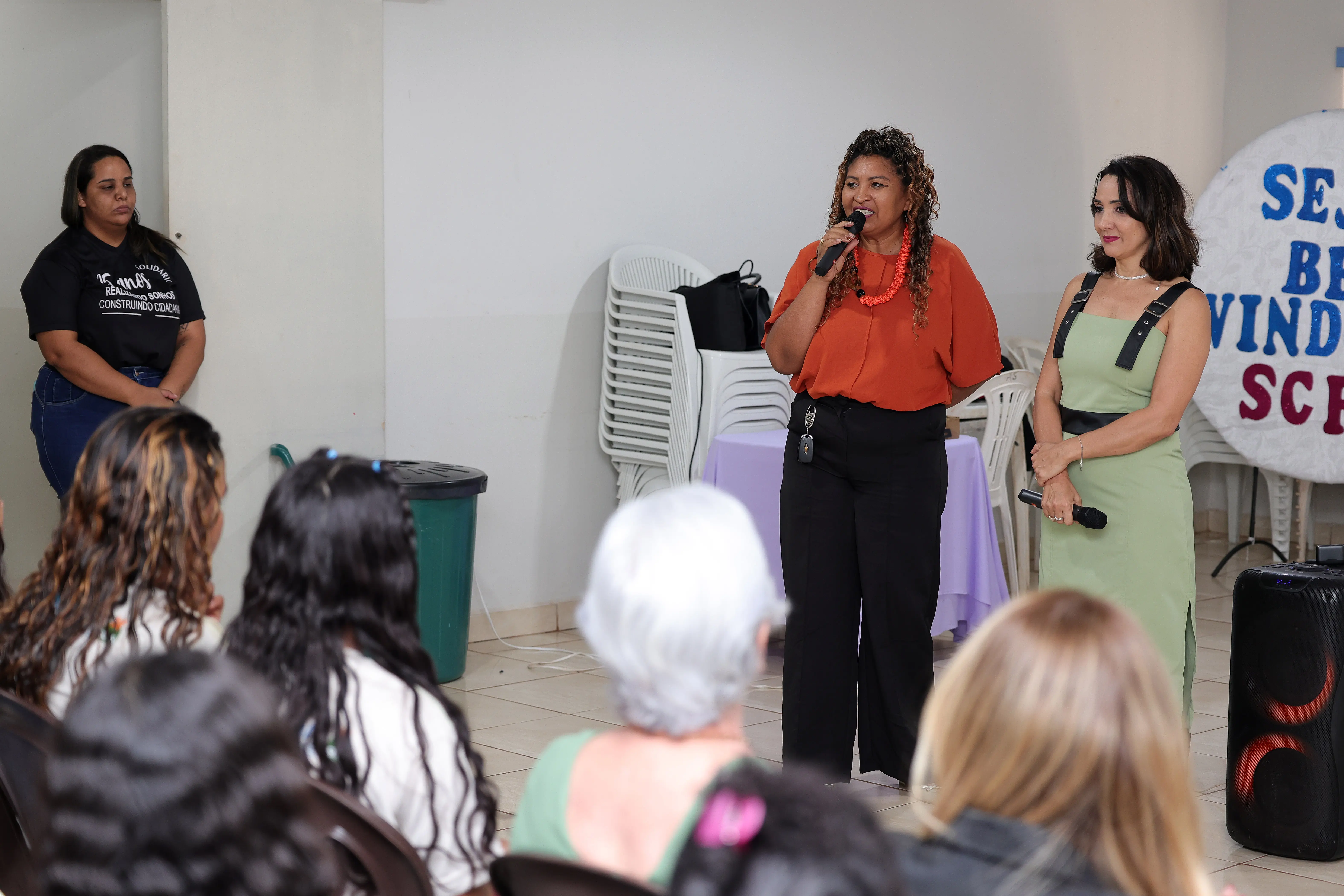 Mulher de blusa terracota fala ao microfone ao lado de outra mulher de vestido verde clara, em pé diante de uma plateia. À esquerda, uma mulher de camiseta preta observa.