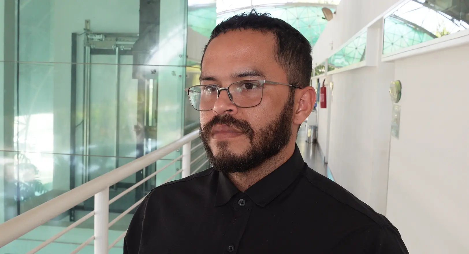 Psicólogo Fábio Souza França é um homem de pele parda, óculos de grau, cabelo curto escuro e barba cheia. Ele veste camisa preta abotoada e olha para a esquerda. Ao fundo, um corredor interno iluminado com divisórias de vidro.