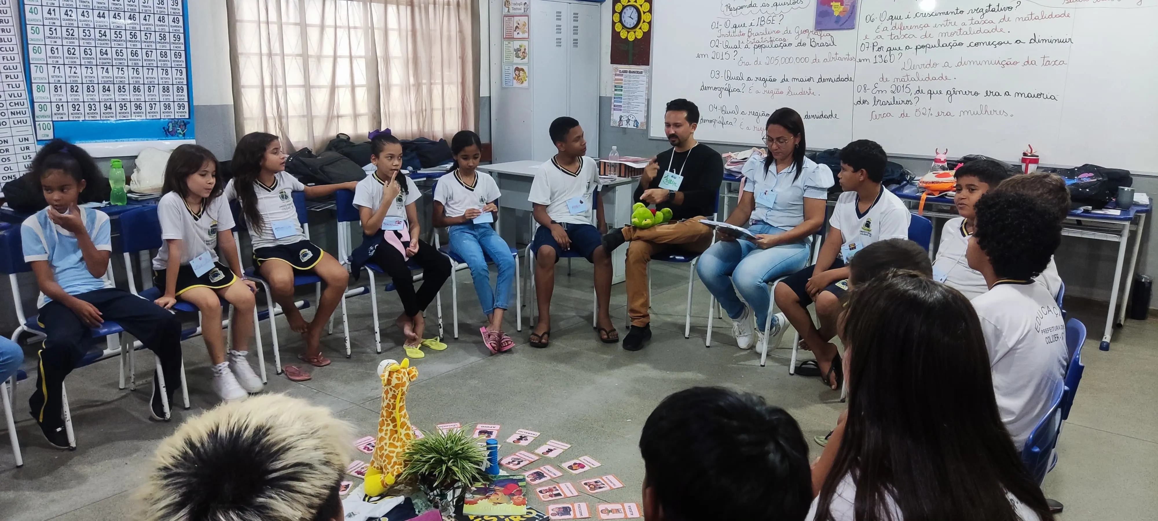 Sentados em círculo, alunos do quinto E da Escola Municipal Atalaia no município de Colíder participam do primeiro círculo de construção de paz. Em volta do objeto de centro que é formado por uma girafa marrom de pelúcia, uma lanterna azul pequena, um livro infantil, uma planta na cor verde e vários