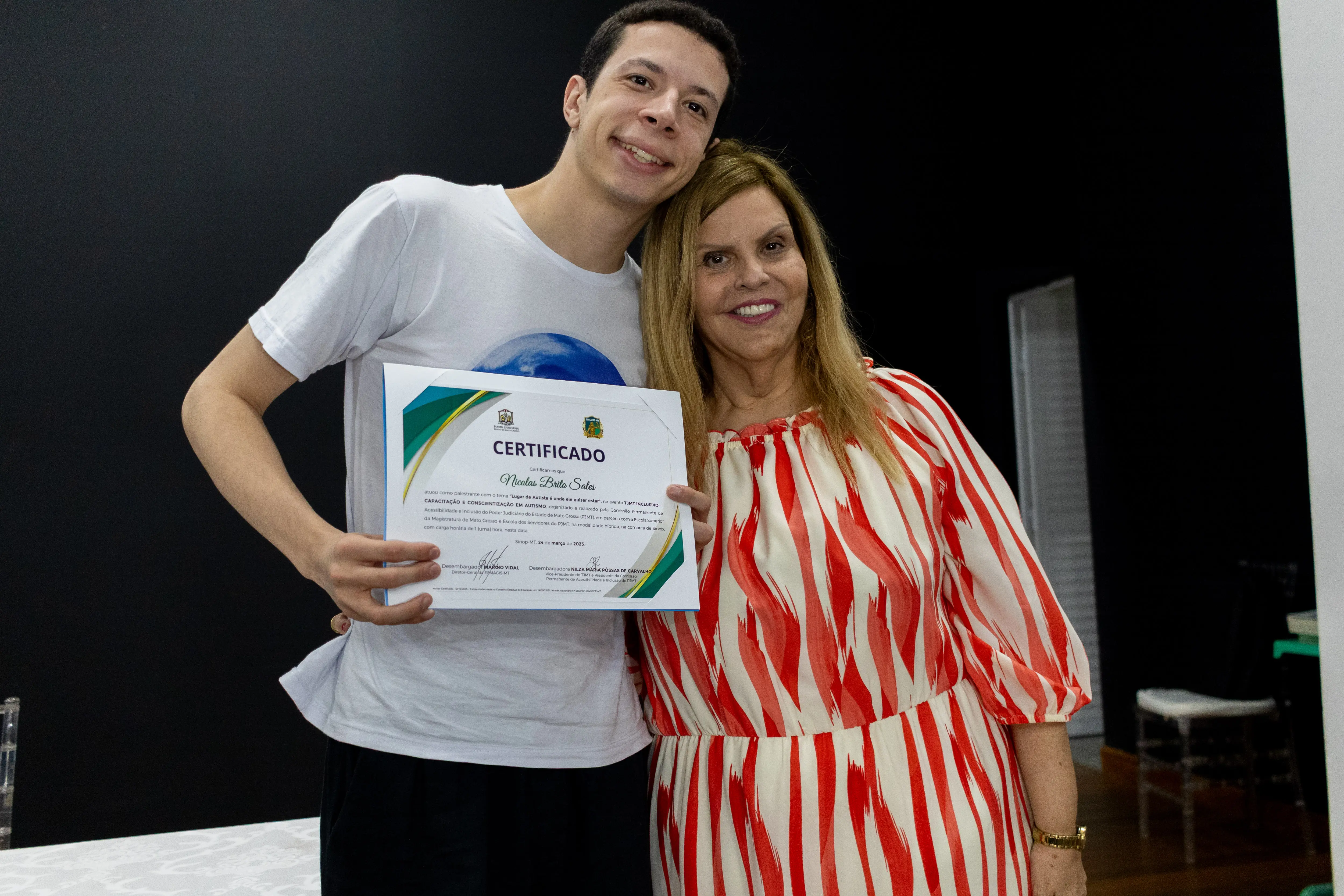 Nicolas abraçado de lado com a desembargadora Nilza durante o TJMT Inclusivo em Sinop. Ele segura um certificado na s mãos