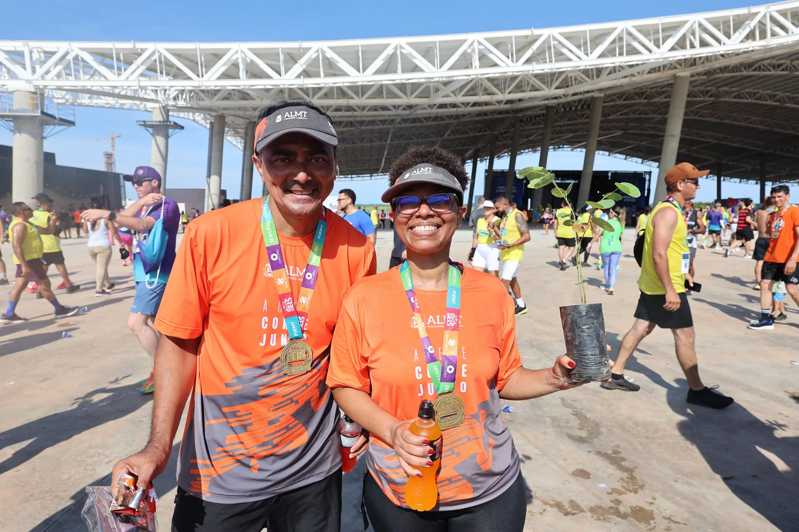 Casal de corredores posa sorridente em espaço externo do evento, vestindo camisetas laranja e medalhas. A mulher segura uma muda de árvore e uma garrafa, enquanto outros atletas caminham ao fundo sob o pavilhão.