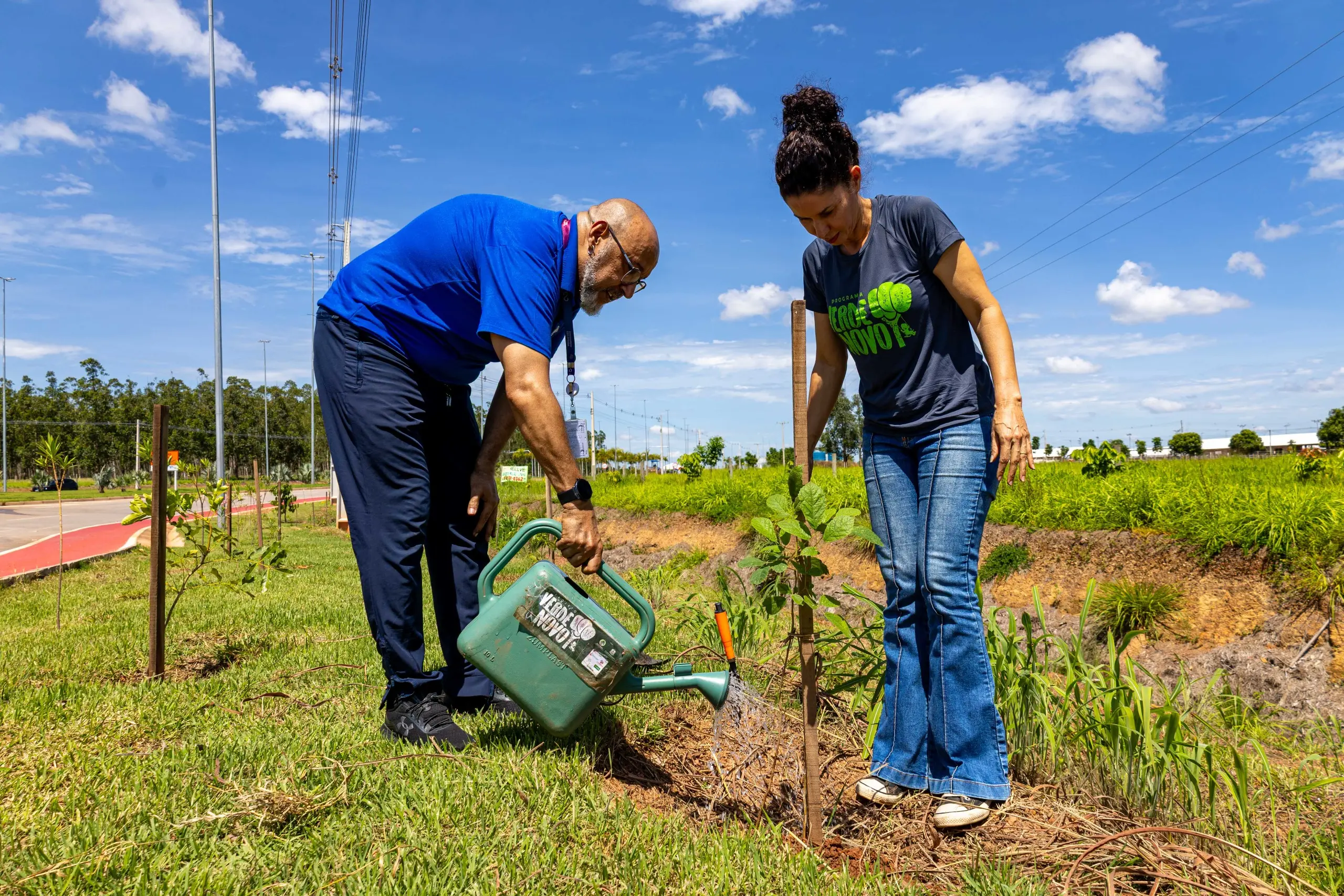 Homem rega a muda recém-plantada enquanto mulher observa o plantio. Ambos usam roupas casuais. A cena ocorre em área verde urbana, com gramado, talude e vegetação ao redor.