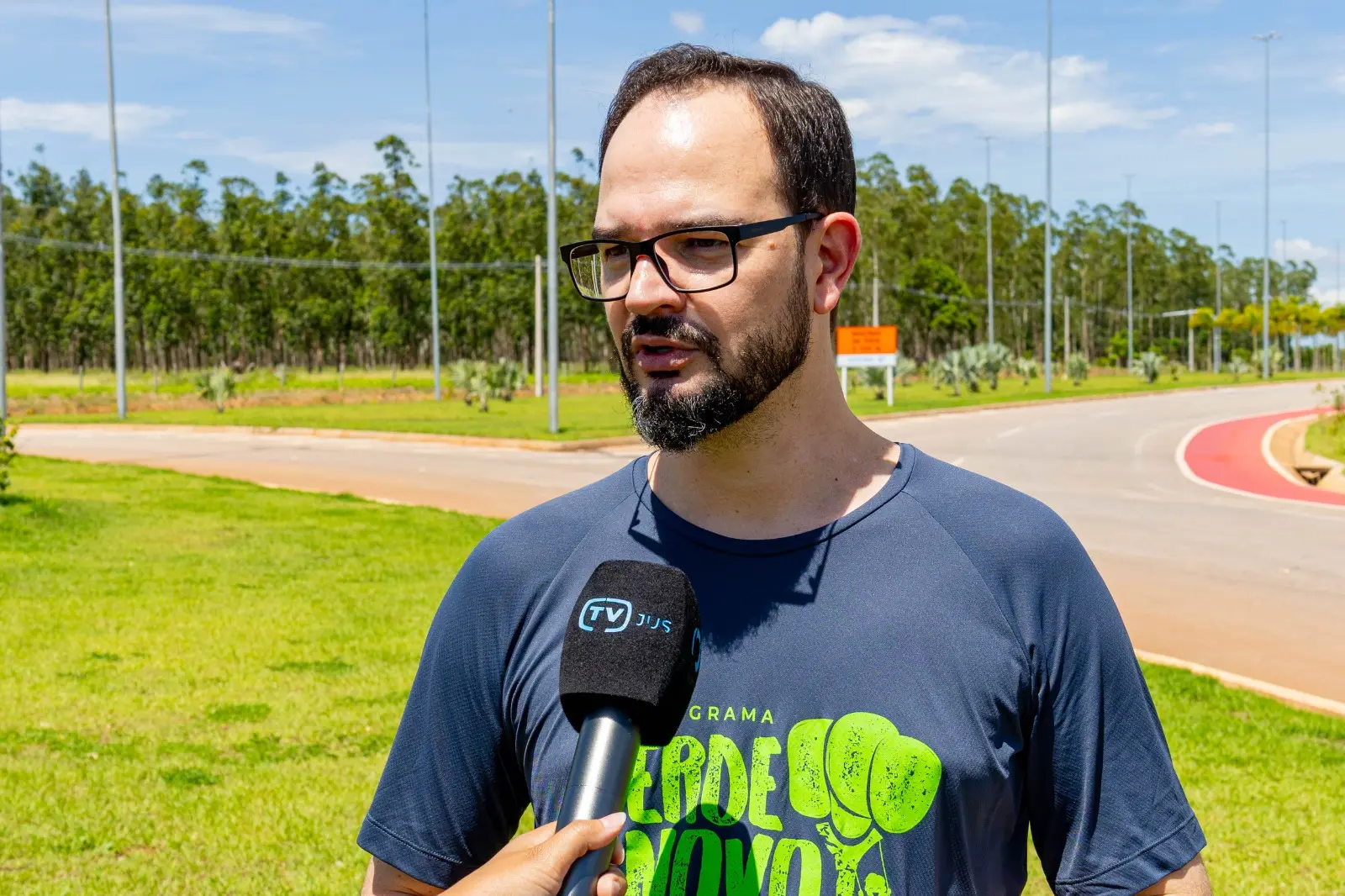 Homem de camiseta do programa Verde Novo concede entrevista ao ar livre. Ele está em pé, diante de área verde com pista, placas e árvores ao fundo, em local amplo e ensolarado.
