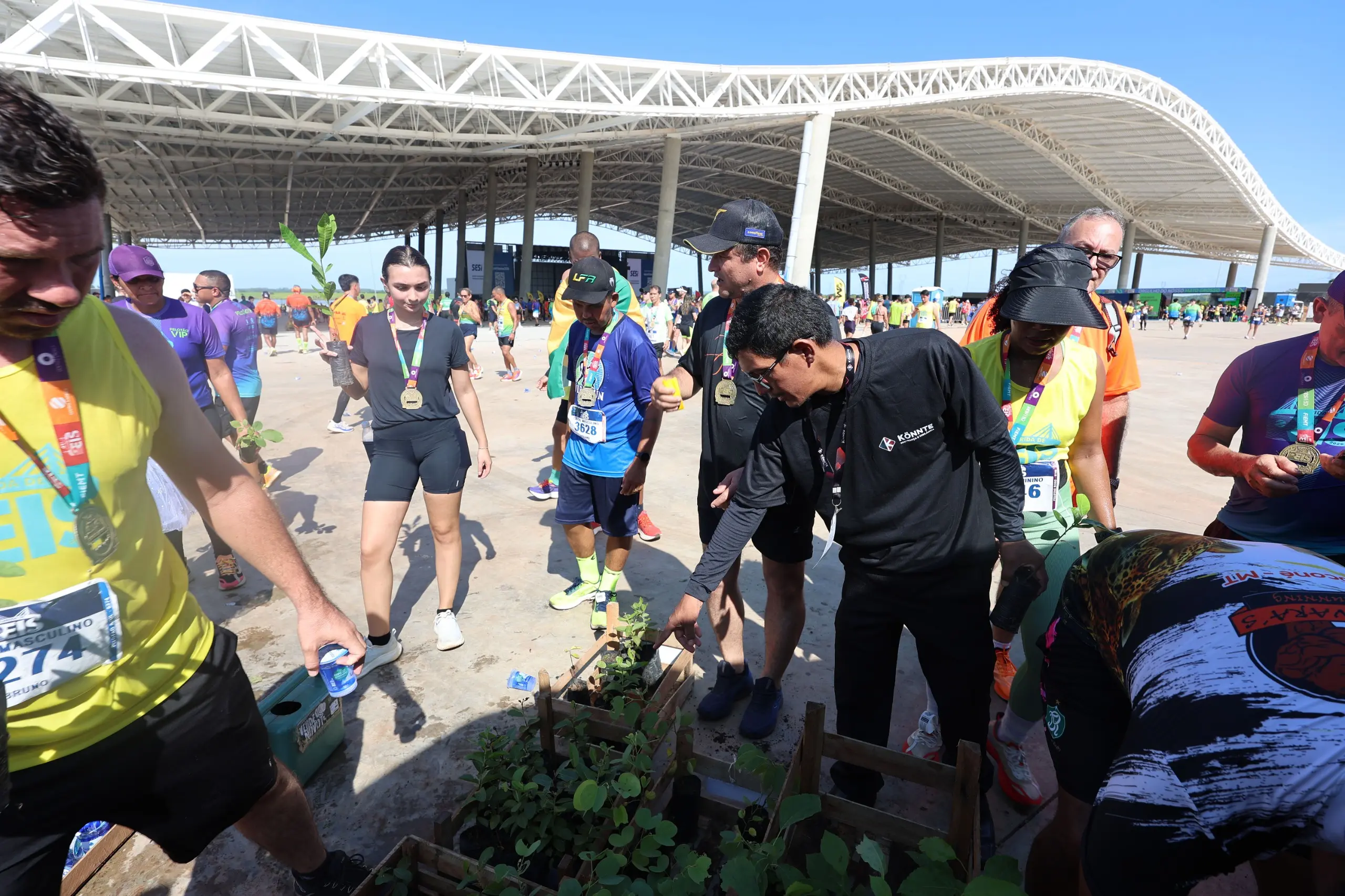 Participantes da corrida se reúnem em torno de caixas de madeira com mudas de árvores. Homens e mulheres, com medalhas e camisetas do evento, escolhem as plantas em espaço amplo, ao lado da estrutura coberta.