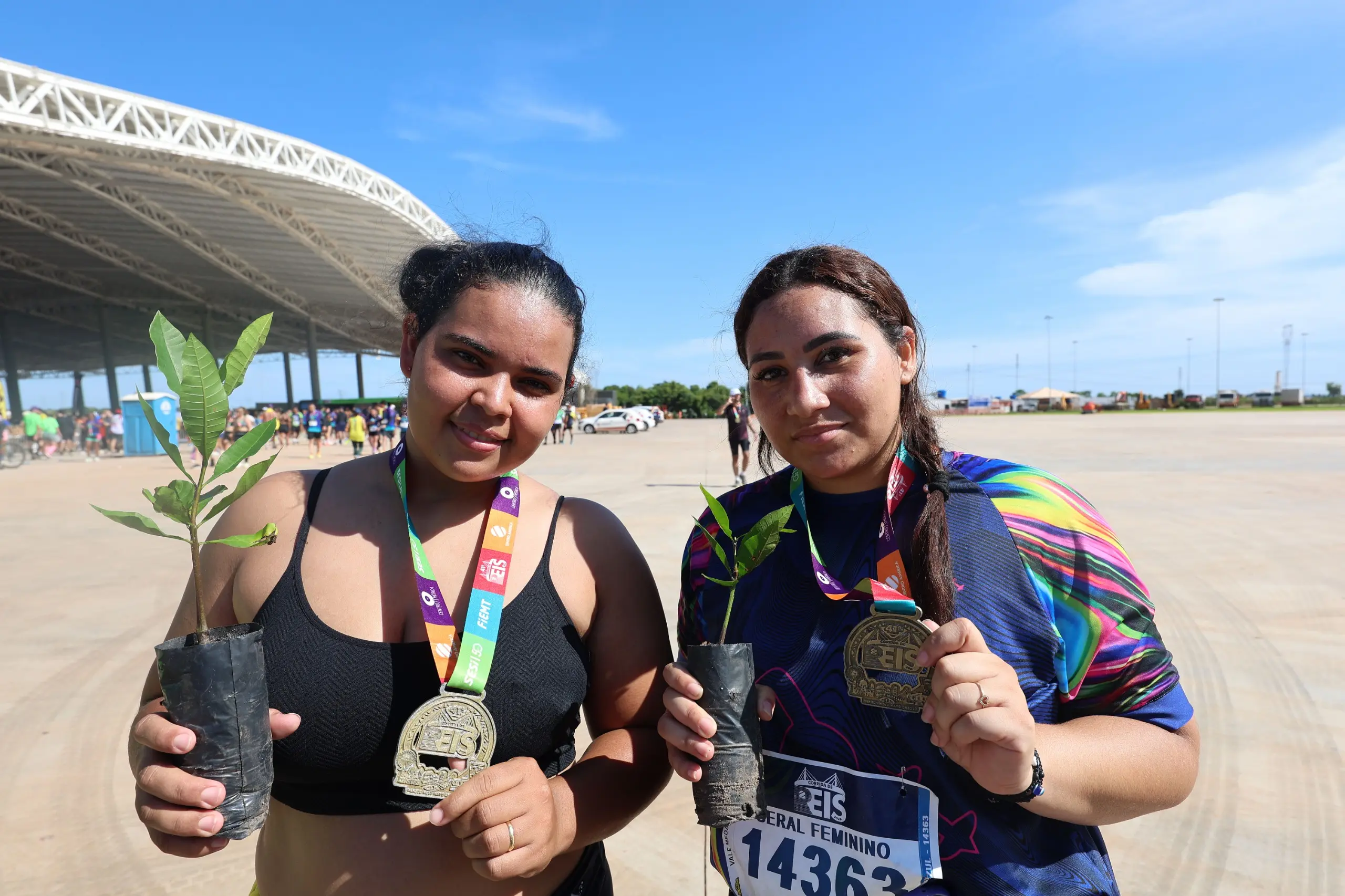 Duas mulheres posam em área externa do evento, usando medalhas da corrida e segurando mudas de árvores em sacos plásticos. Ao fundo, pavilhão coberto, céu azul e outros participantes circulam após a prova.