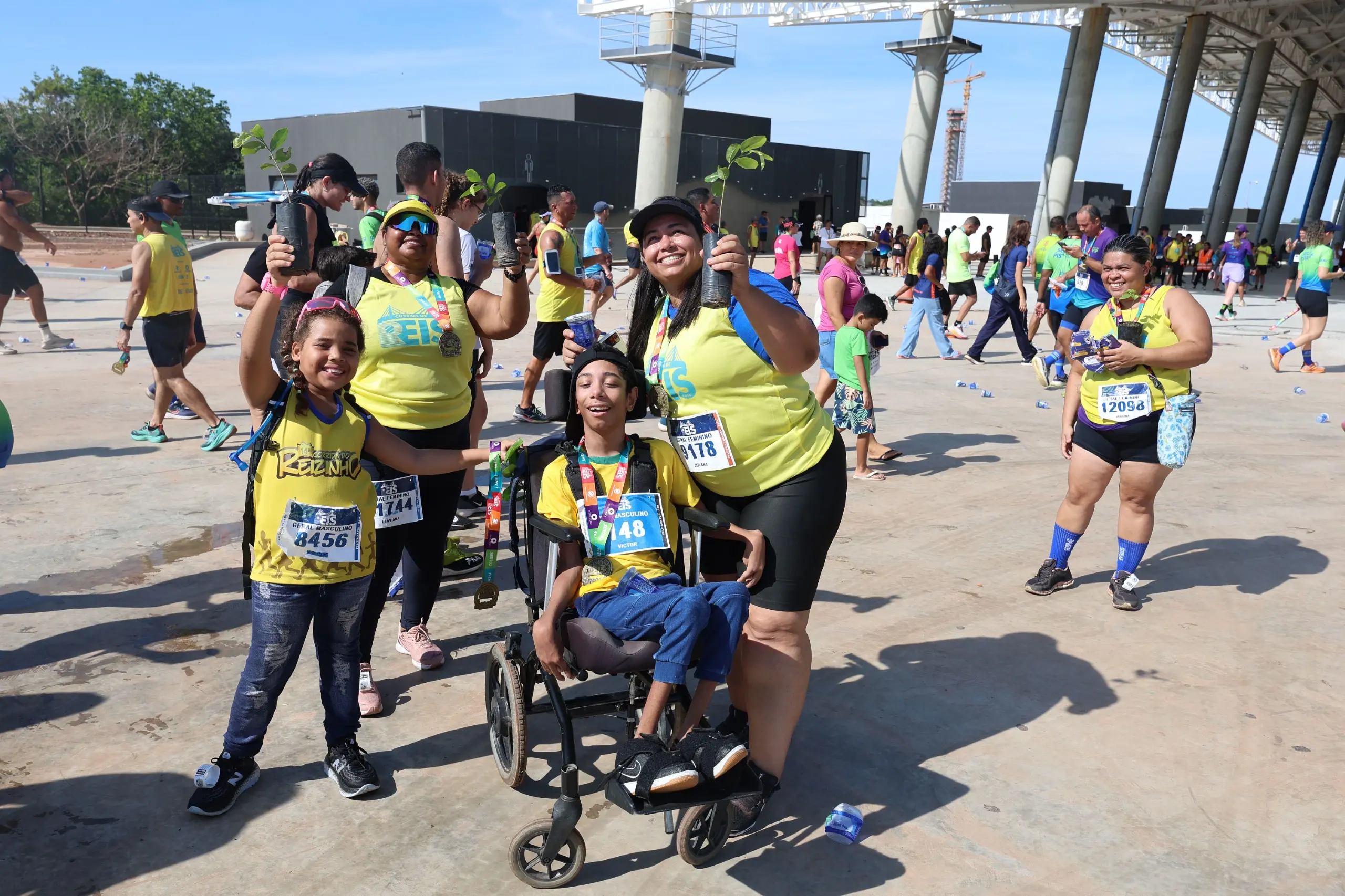 Grupo diverso celebra após a corrida, incluindo crianças e um participante em cadeira de rodas. Todos usam camisetas amarelas, medalhas e seguram mudas de árvores, em área aberta com grande movimentação ao fundo.