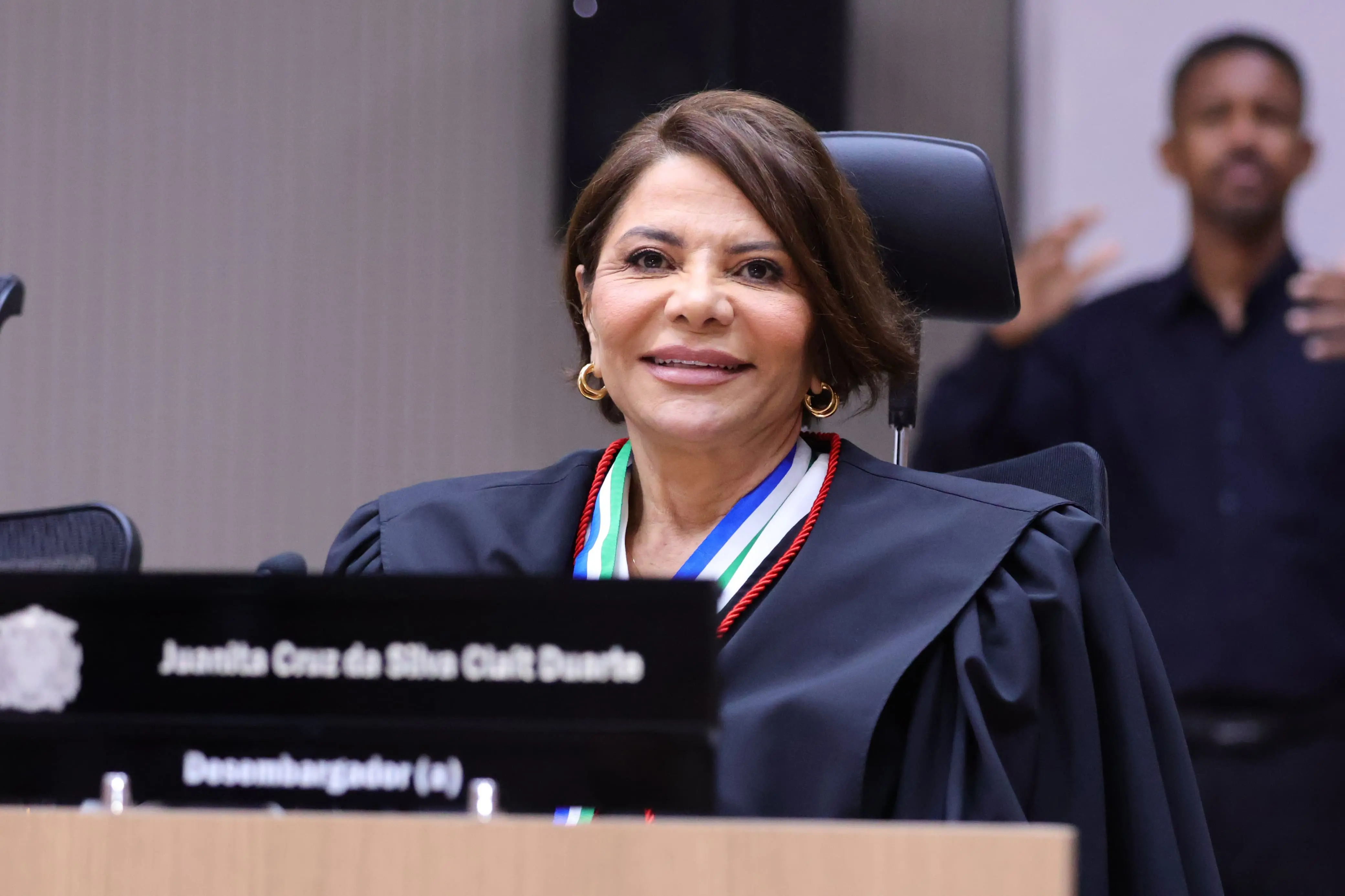 Imagem de uma mulher sorridente, vestida com toga e medalhas no pescoço, sentada à mesa com placa identificando-a como desembargadora Juanita Cruz da Silva Clait Duarte. Ao fundo, um intérprete de Libras aparece em atuação.