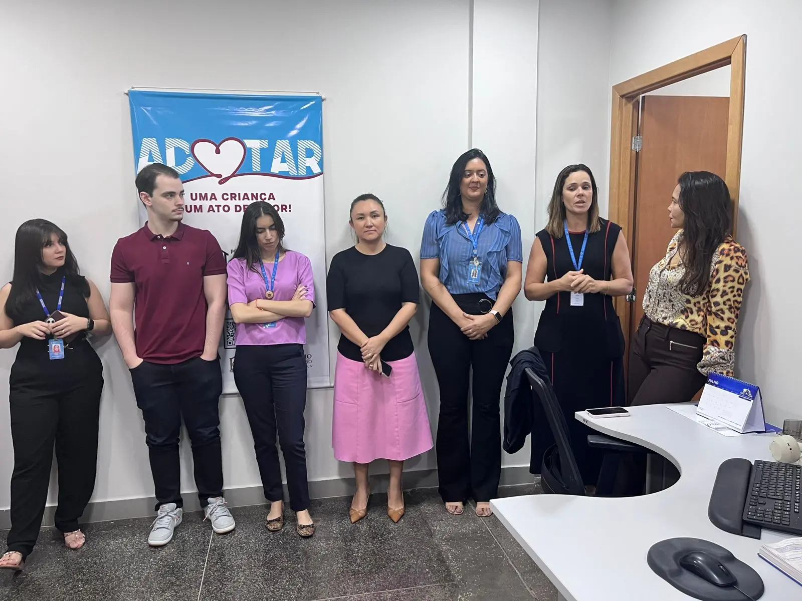 mostra um grupo de seis pessoas em pé, enfileiradas contra a parede. Atrás do grupo, há uma faixa na parede com a palavra “adotar” e um símbolo de coração.