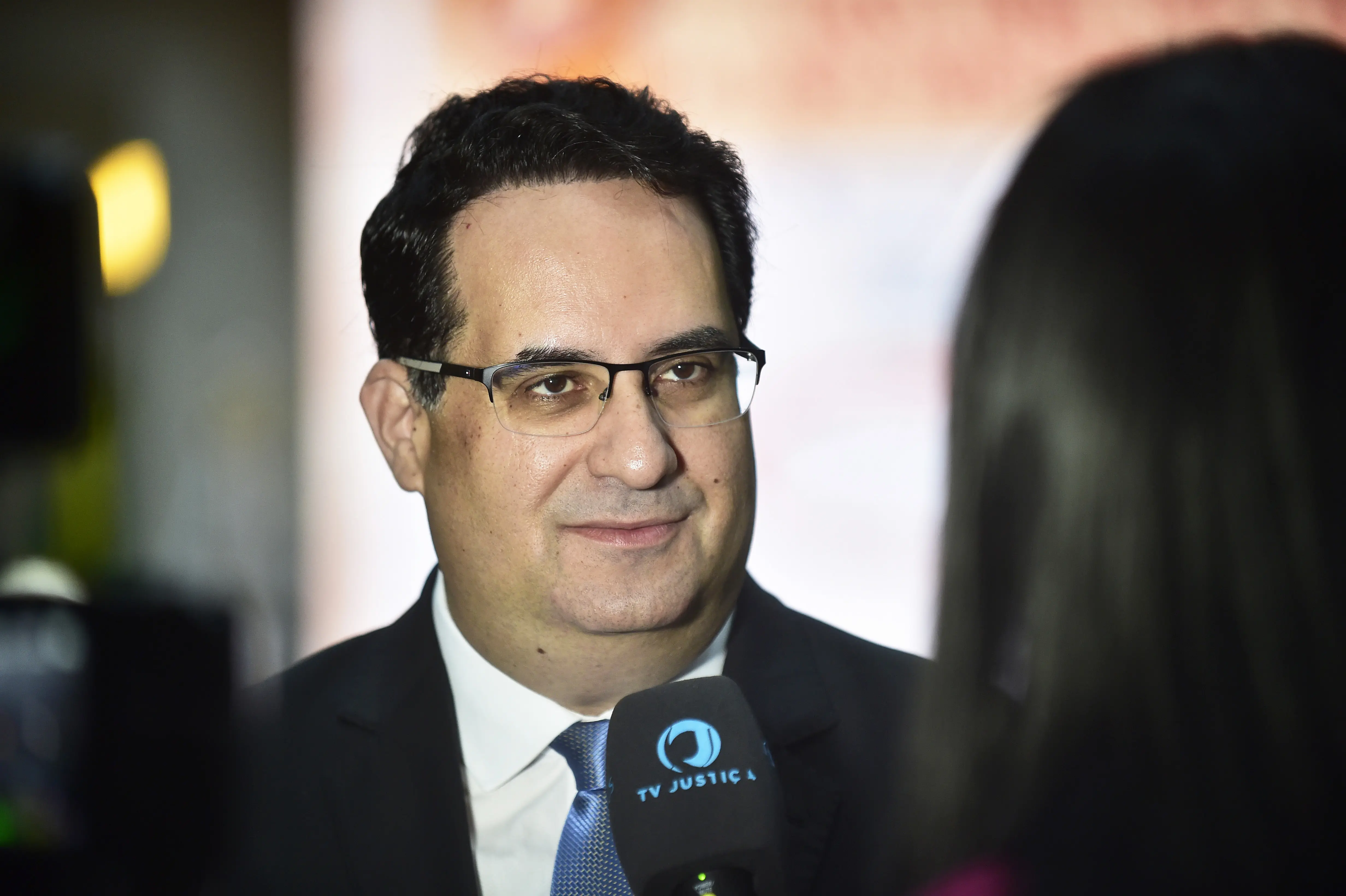 Juiz Túlio Duailibi Alves Souza tem pele clara e cabelo escuro, com óculos, terno e gravata azul. Ele sorri levemente enquanto segura um microfone preto com o logo "TV JUSTIC A". Uma pessoa está de costas no canto direito.