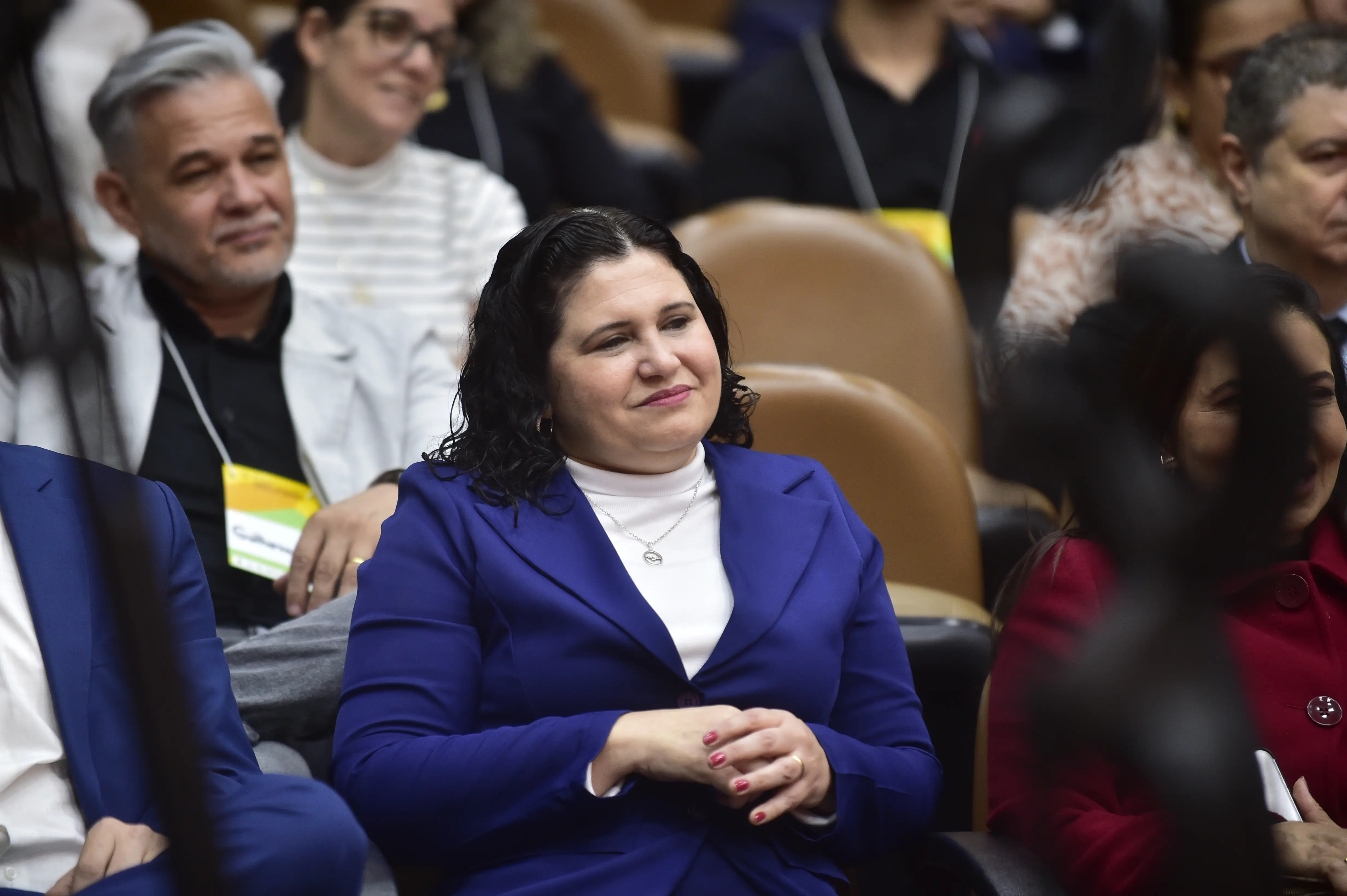 Uma mulher de cabelos escuros e ondulados, usando um blazer azul sobre uma blusa branca de gola alta e um colar, está sentada em um ambiente com outras pessoas borradas ao fundo, sorrindo gentilmente e com as mãos cruzadas.