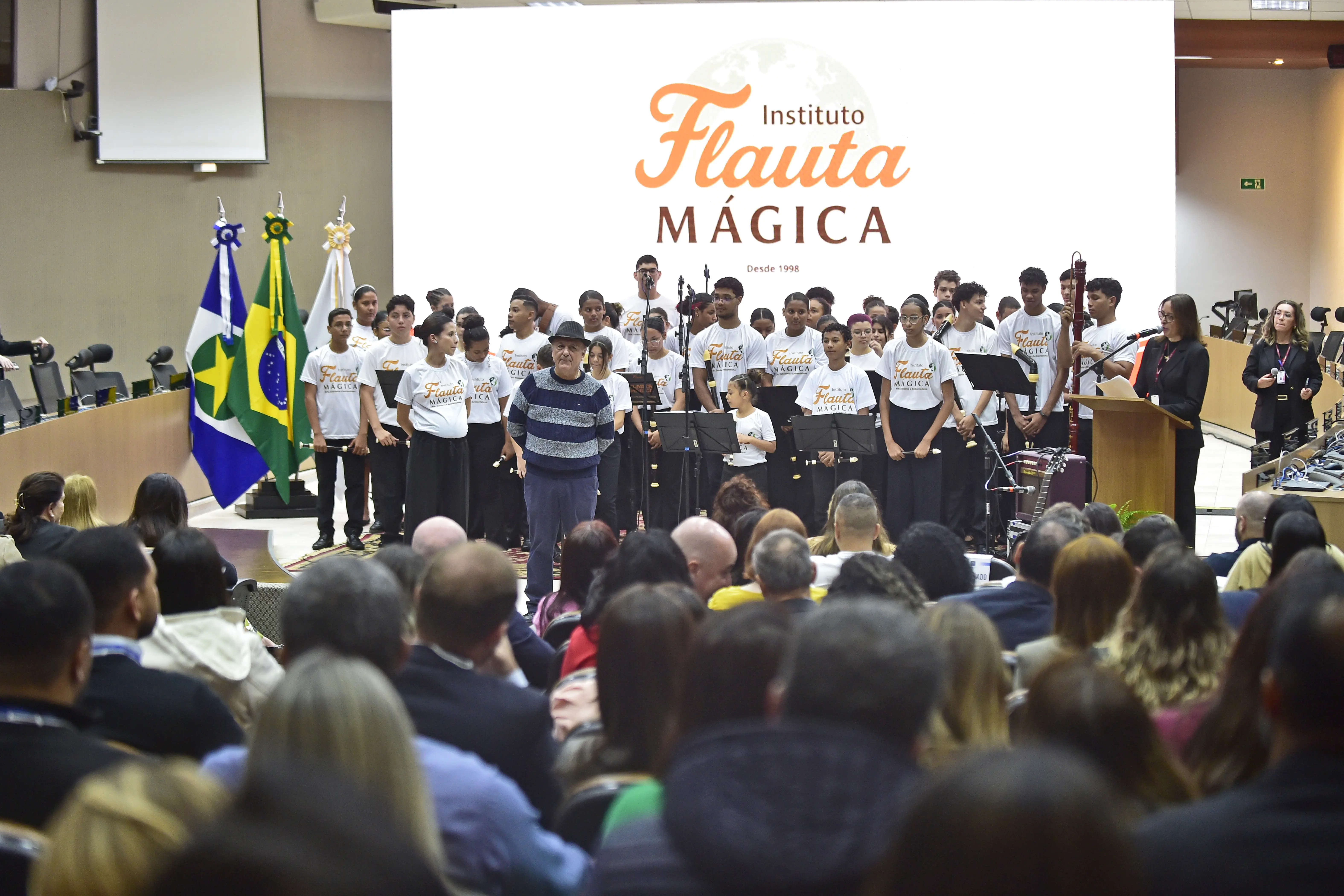 Palco com um grande telão branco exibindo "Instituto Flauta Mágica". Um grupo de jovens em camisetas brancas está organizado no palco, com um homem de chapéu à frente, ao centro. À esquerda, bandeiras. Uma plateia observa de costas.