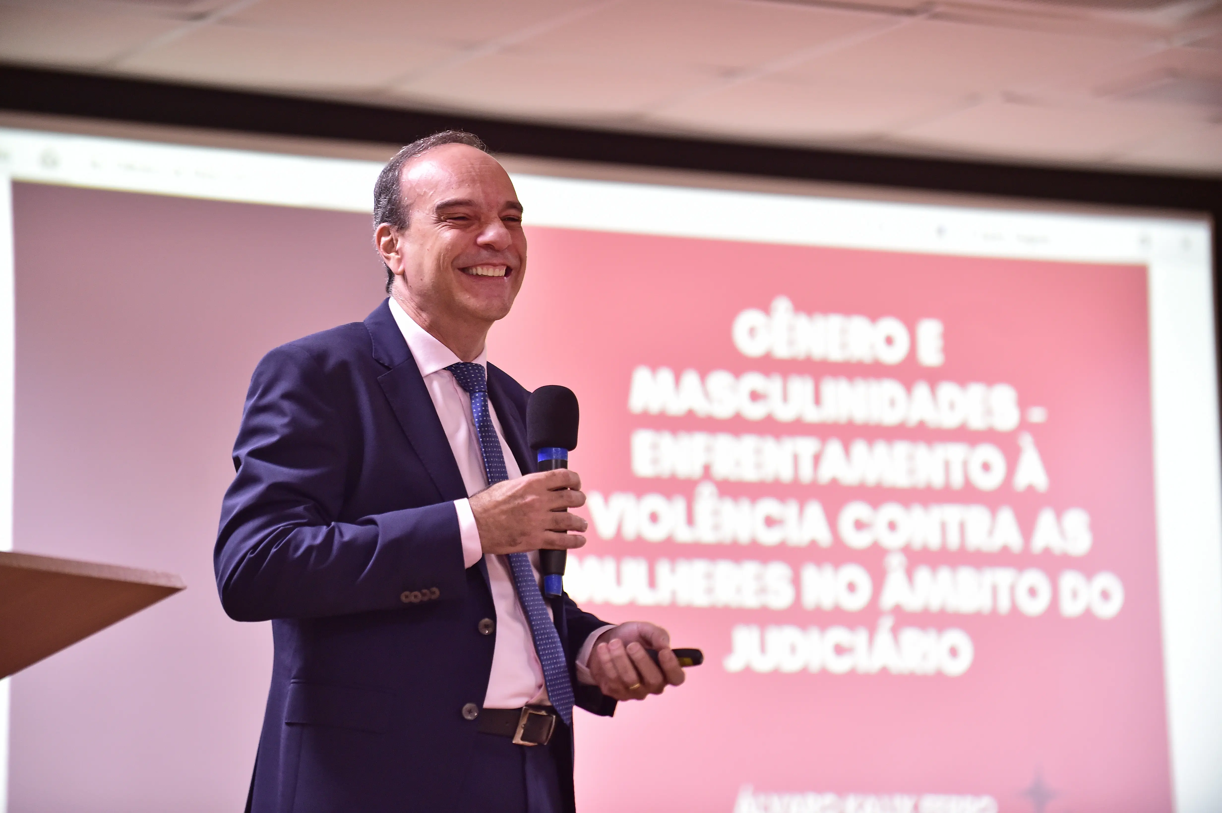 Foto do desembargador do TJ de Rondônia, Álvaro Kálix, proferindo palestra e sorrindo. Ele é um homem branco, alto, magro, usando camisa rosa clara, terno e gravata azuis.