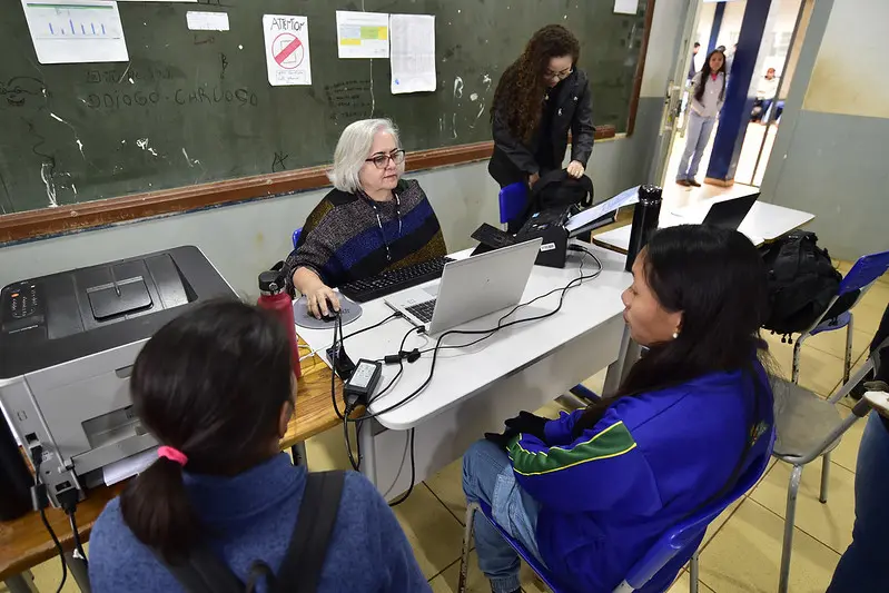 Sala de aula onde uma servidora da Receita Federal atende a duas mulheres. ela manuseia o notebook, enquanto as mulheres aguardam.