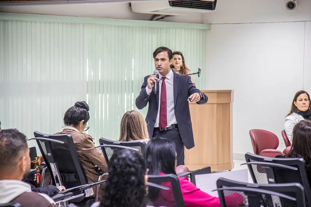 Homem de terno escuro e gravata vermelha fala em um microfone, gesticulando para o público. Uma mulher loira atrás de um púlpito o observa. À direita, outra mulher sentada na plateia e várias pessoas de costas.   Fontes