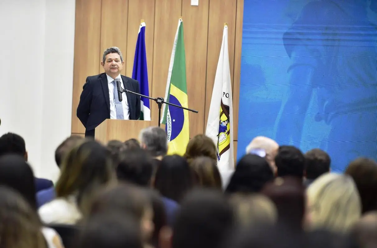 Homem de terno, com cabelo grisalho, discursa em púlpito de madeira. Atrás dele, três bandeiras: azul, do Brasil e branca. Uma plateia diversificada o ouve atentamente, com foco em suas expressões e gestos