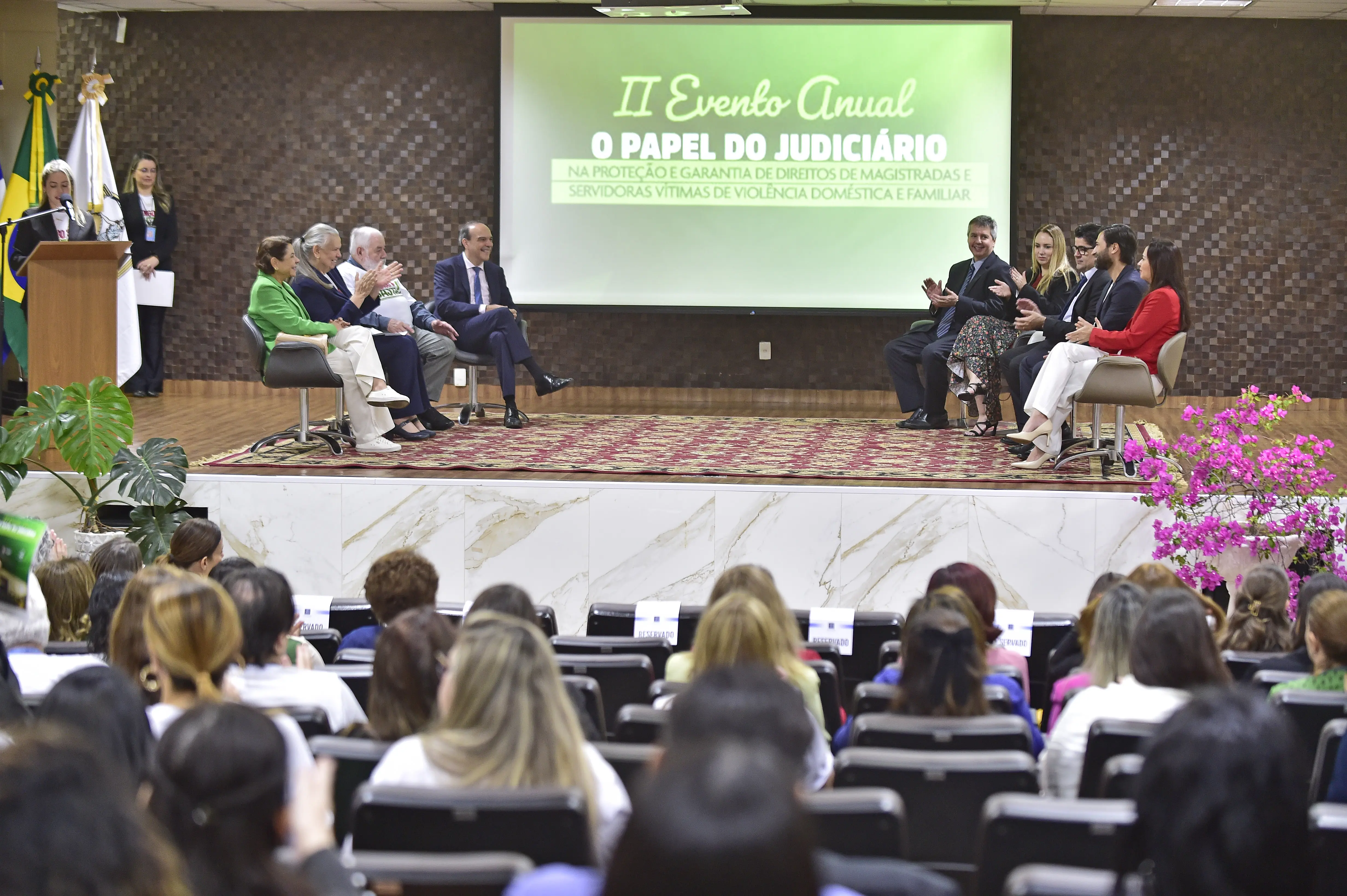 Foto que mostra o auditório do TJMT repleto de pessoas na plateia e autoridades sentadas em poltronas no palco. No telão aparece a arte do evento na cor verde.