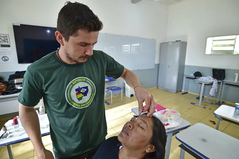 Profissional faz procedimento de sobrancelha de henna em mulher, que está sentada com os olhos fechados, em uma sala de aula.