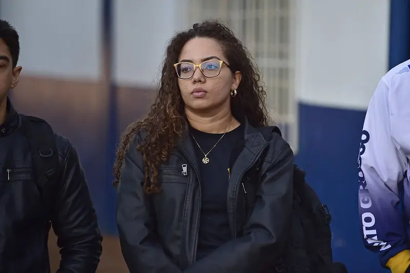 Policial federal em pé. Ela é uma mulher jovem, de cabelos longos e cacheados, usando óculos de grau e jaqueta de couro preta.