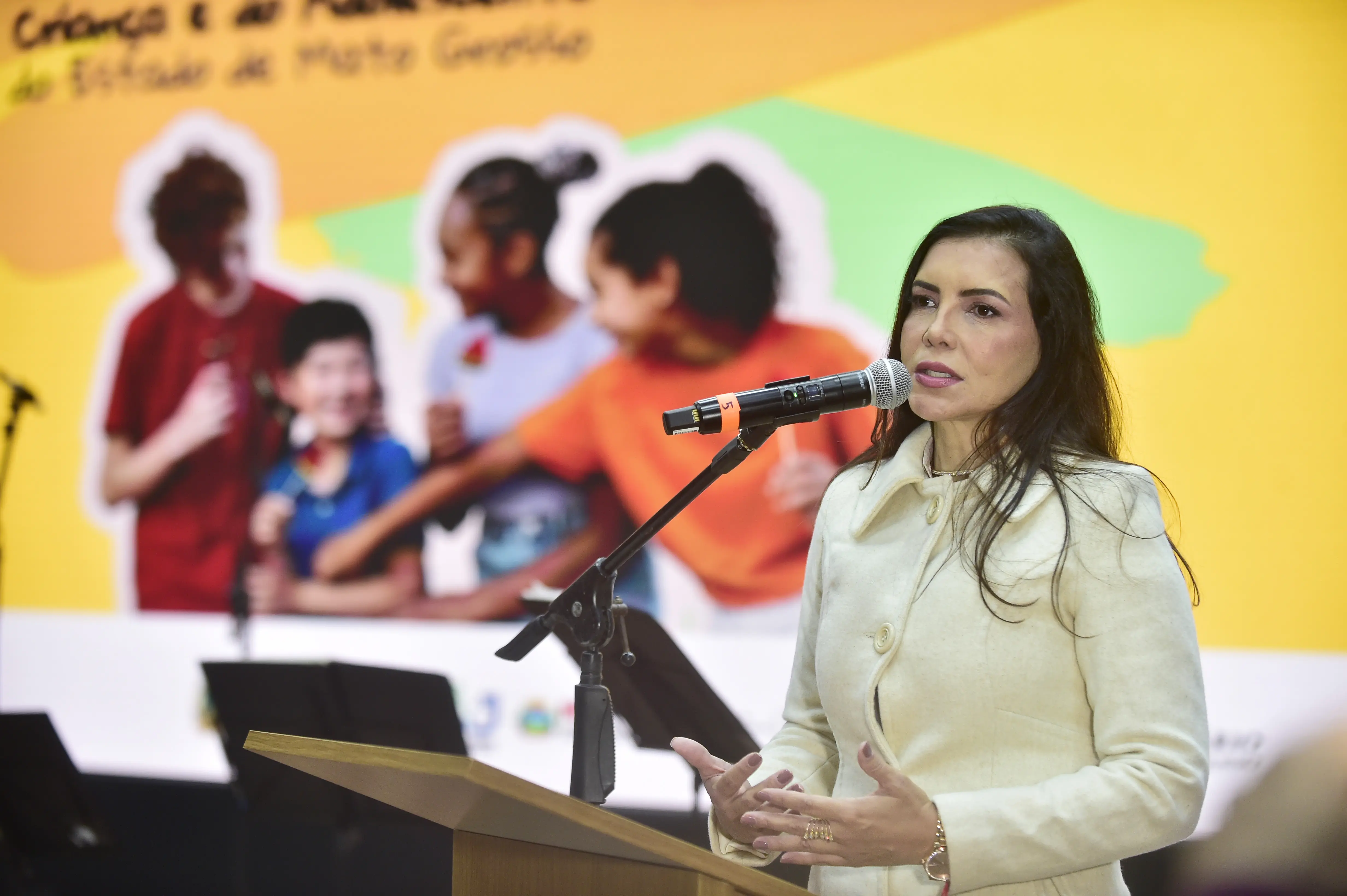 Juíza Anna Paula Gomes de Freitas tem cabelos escuros e compridos, veste um casaco creme. Ela está em pé, atrás de um púlpito de madeira escura, falando em um microfone preto com uma gola amarela. O fundo é vibrante, com imagens de pessoas.